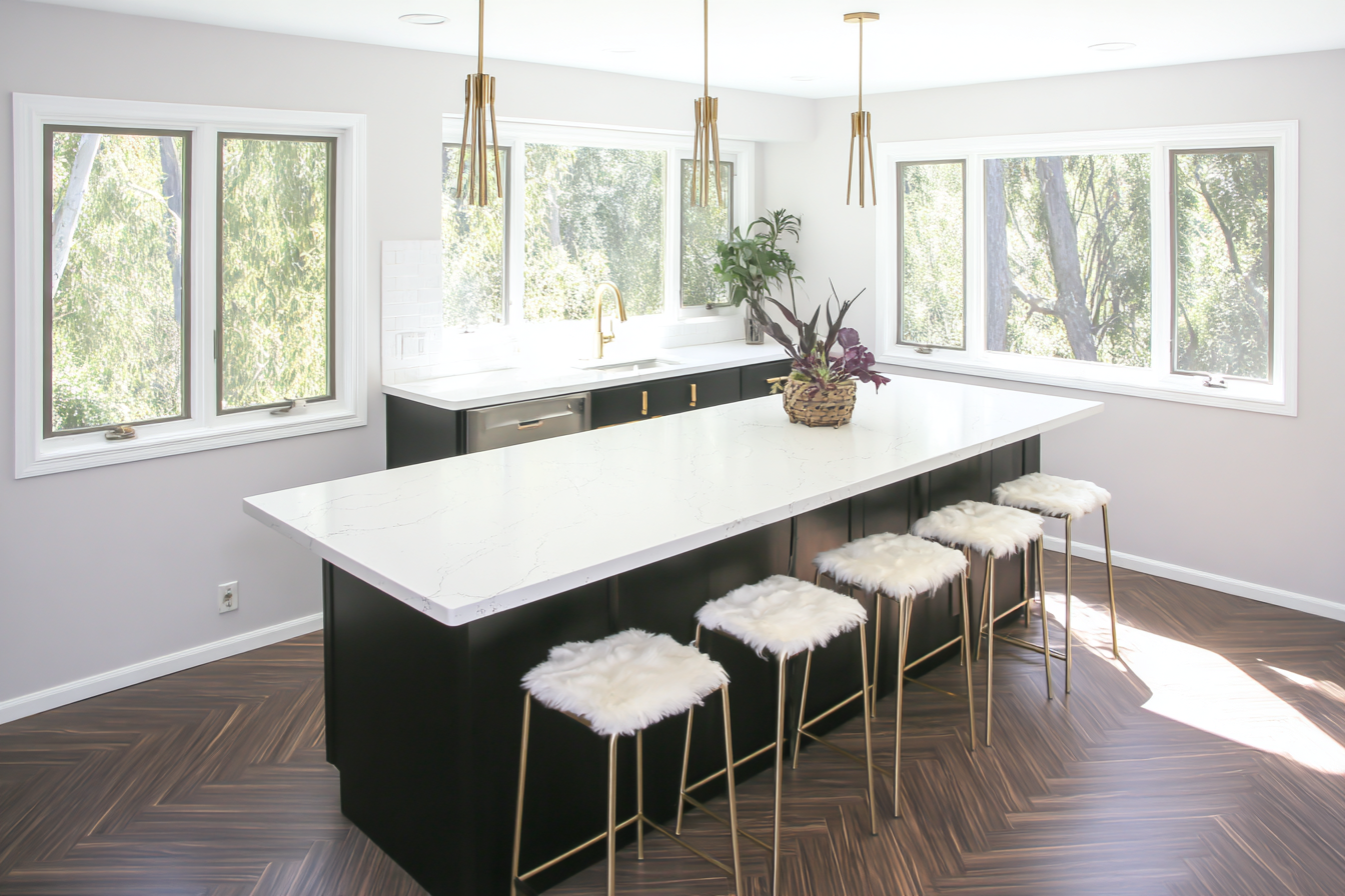 Cozinha moderna iluminada com grandes janelas, bancada longa de mármore branco e banquetas com assento felpudo. Iluminação natural realça o piso em espinha de peixe, criando um ambiente sofisticado e acolhedor.