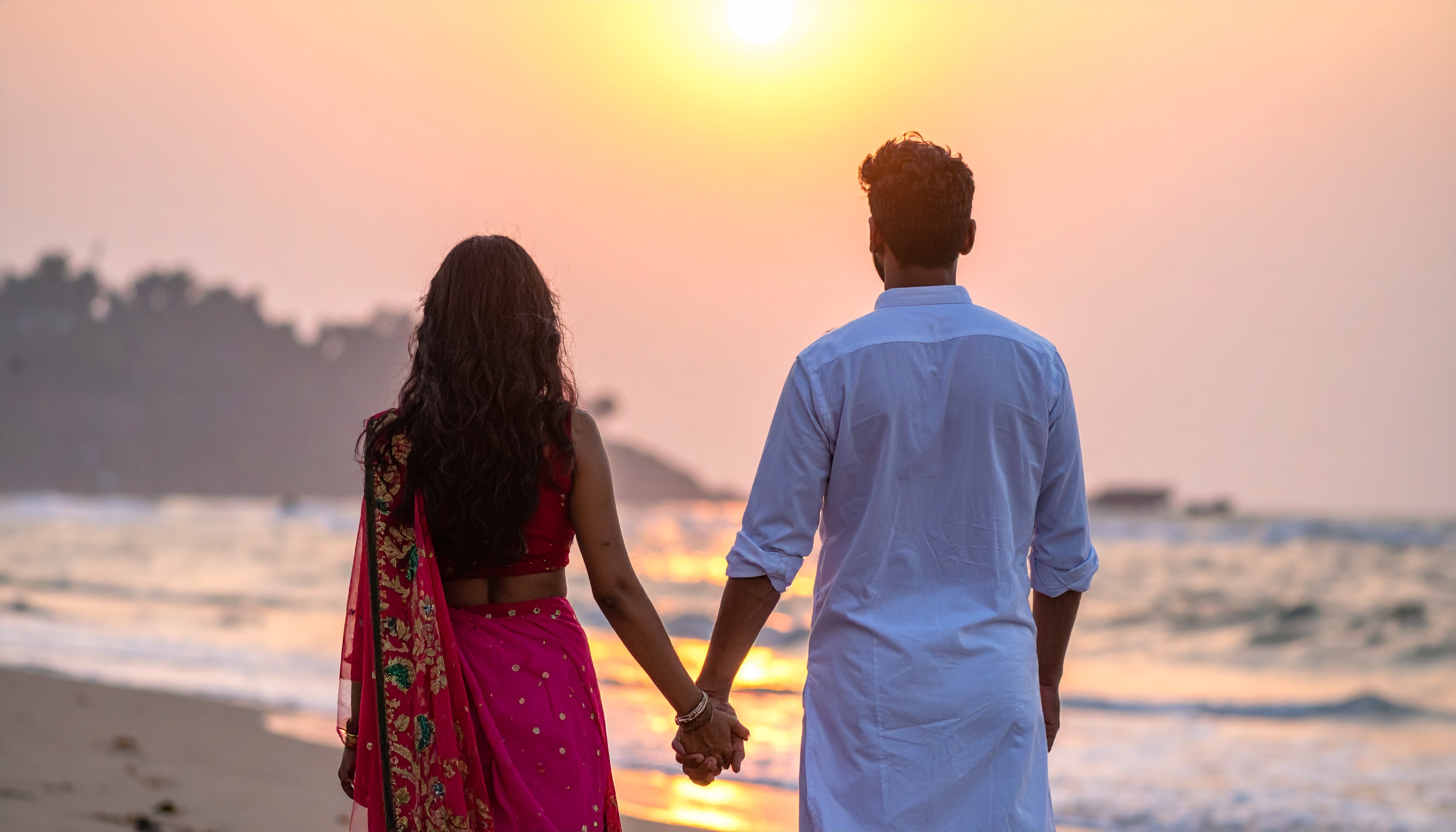 A couple stands hand in hand on a beach at sunset, creating a romantic scene