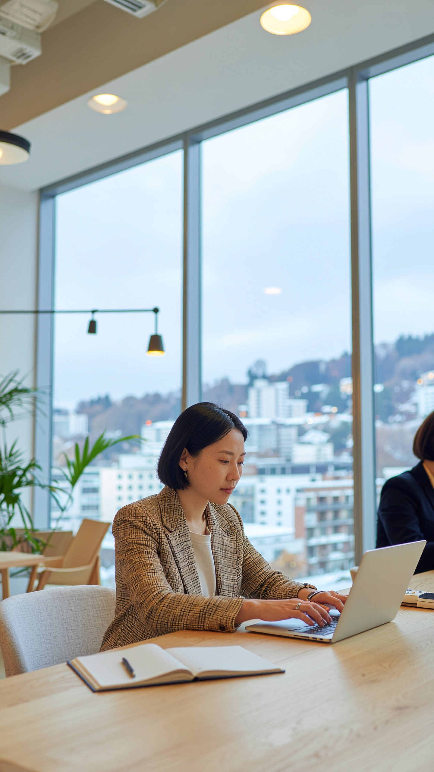 A woman in a modern office works on a laptop by large windows