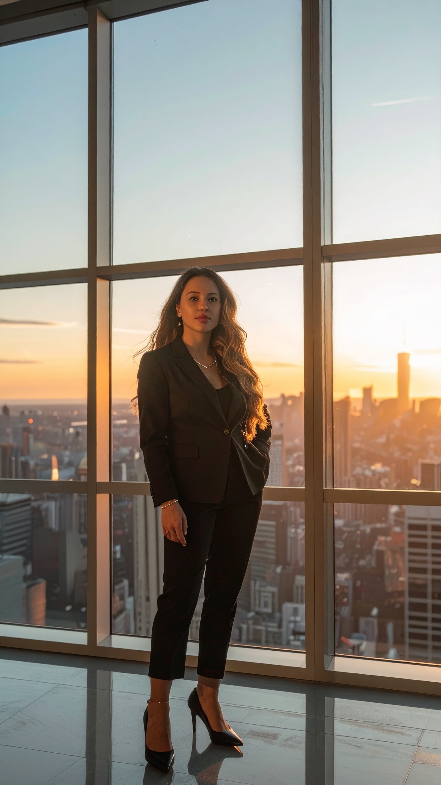 Mulher de terno preto em um arranha-céu moderno, capturada ao pôr do sol com vista urbana, transmitindo elegância e poder no ambiente corporativo.