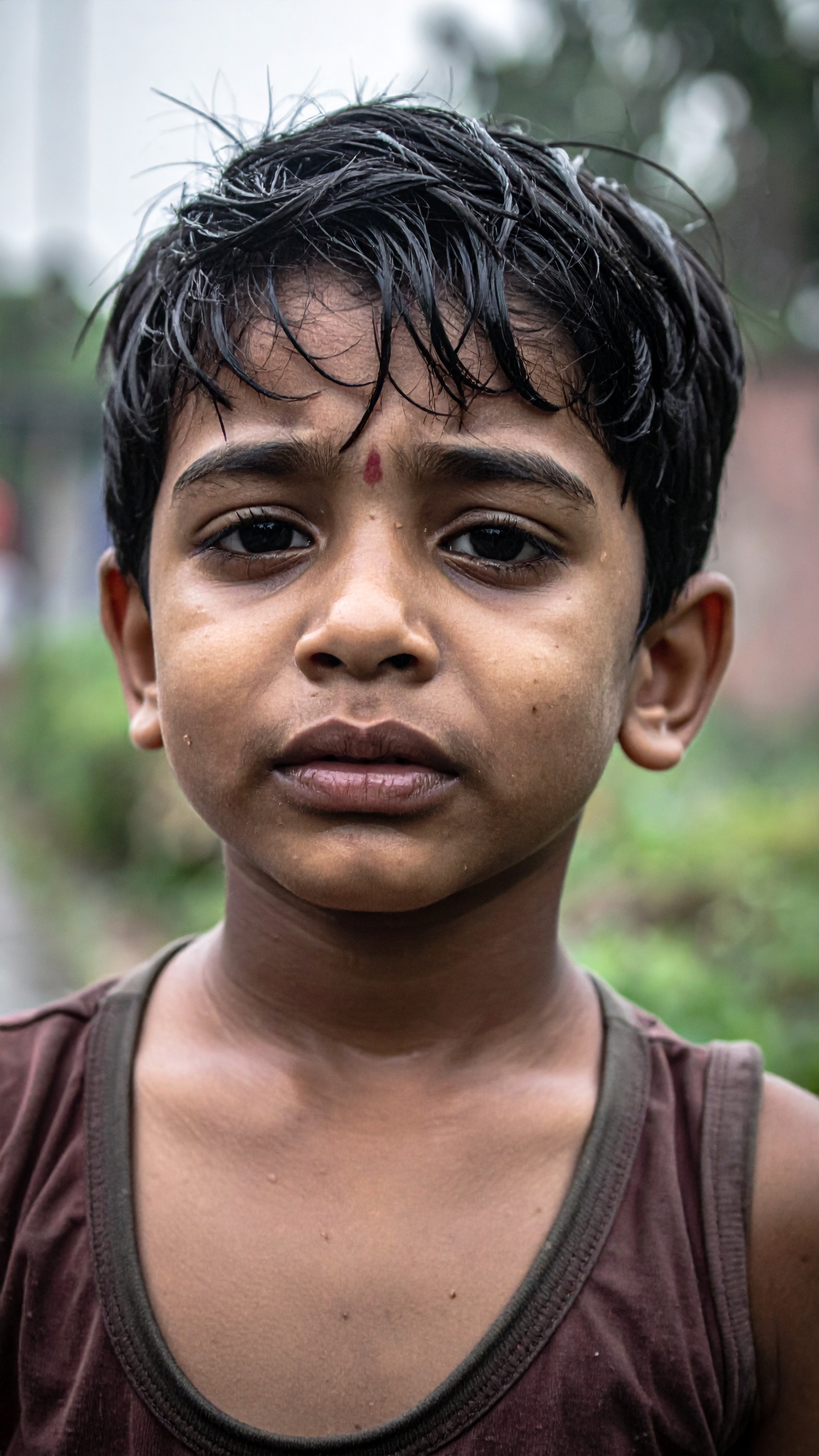 A young boy with a contemplative expression stands outdoors, his hair slightly wet