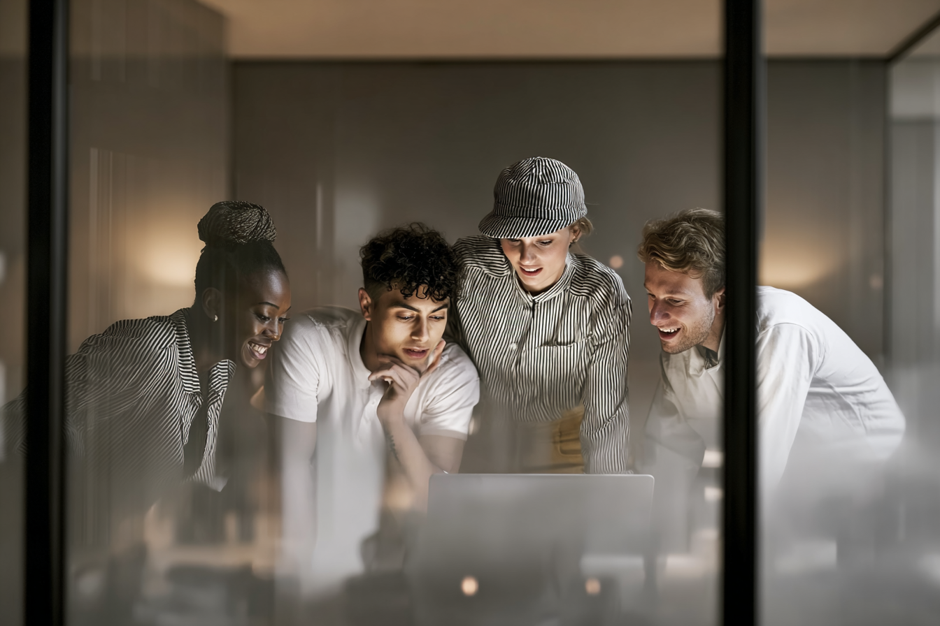 Quatro jovens adultos colaboram entusiasmados em um projeto em frente a um laptop, em um escritório moderno e iluminado, transmitindo criatividade e trabalho em equipe.
