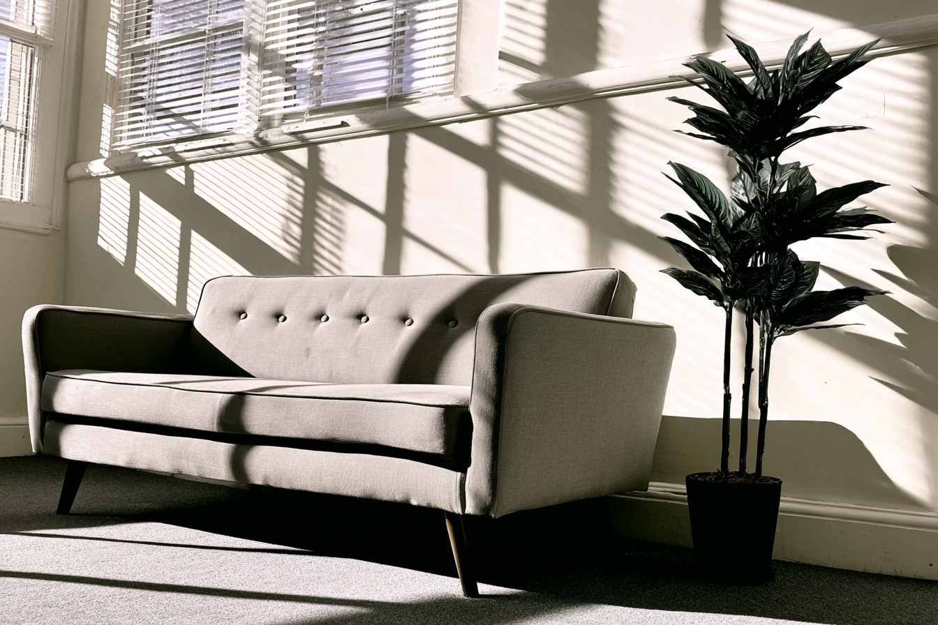 Sofá cinza elegante em sala iluminada por luz do sol filtrada através de persianas, criando sombras geométricas na parede. Planta decorativa ao lado adiciona contraste com suas folhas verdes. Composição minimalista e contemporânea com foco no design e no jogo de luz e sombra.