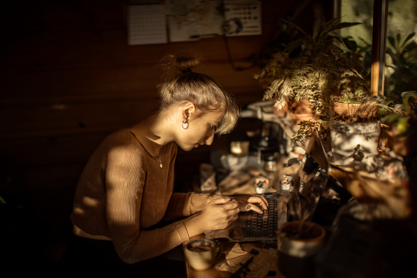 Jovem mulher concentrada em seu laptop em um ambiente aconchegante, com luz suave e plantas ao fundo, ideal para trabalho remoto ou estudos. A mesa está decorada com itens de escritório e uma xícara de café.