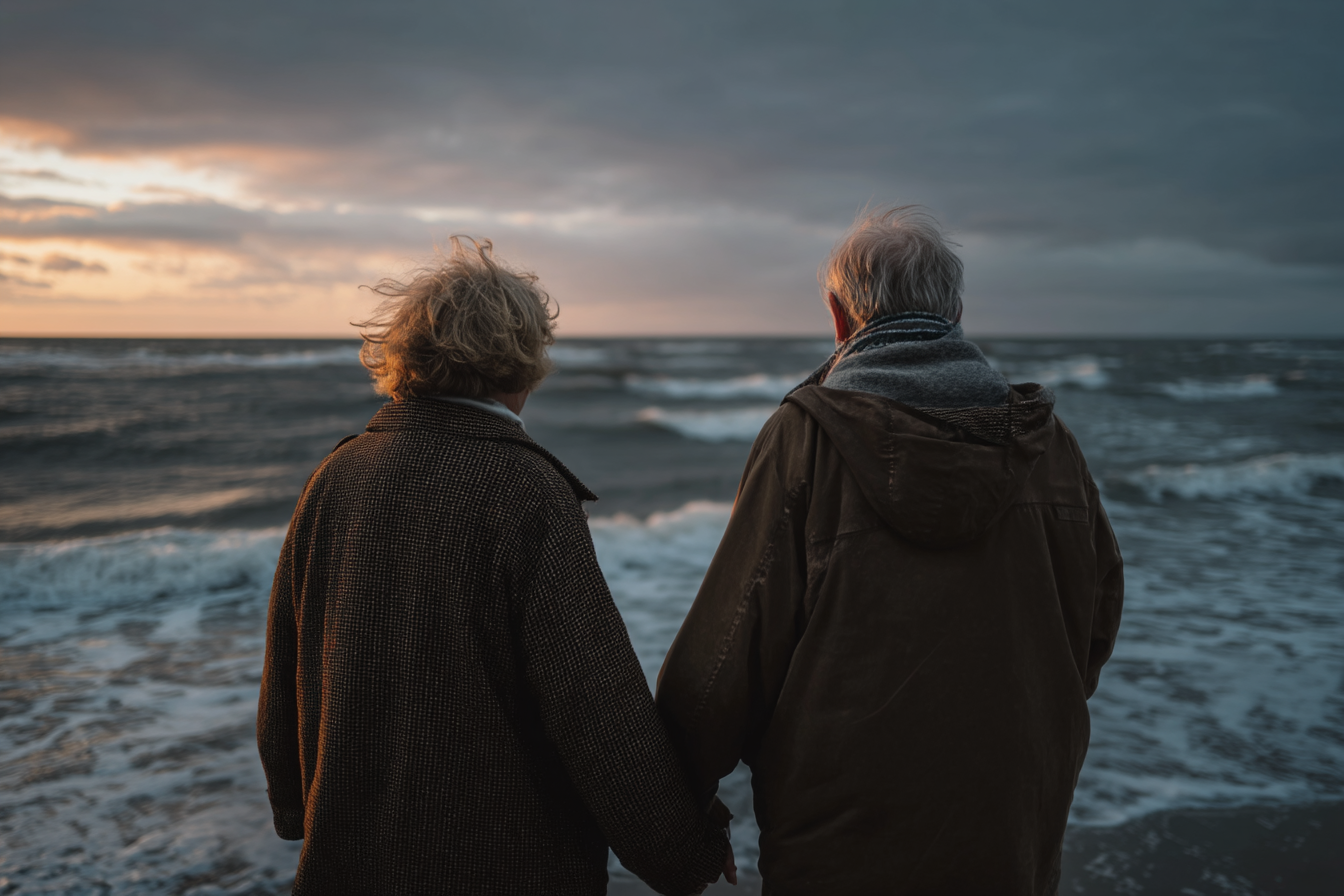 Um casal de idosos de mãos dadas, observando o pôr do sol na praia, com ondas do mar ao fundo e um céu nublado. A cena transmite um sentimento de amor e tranquilidade, ideal para temas de relacionamentos e aposentadoria.