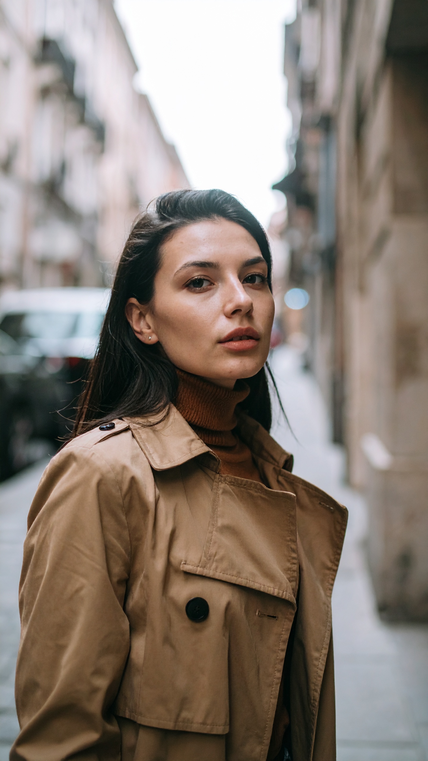 Uma mulher vestindo um casaco bege caminha por uma rua urbana estreita. O casaco é ideal para climas outonais, proporcionando estilo e conforto. Seus botões pretos contrastam elegantemente com o tecido claro, conferindo um toque clássico. Este tipo de vestuário é perfeito para passeios em cidades durante dias frescos e nublados. A versatilidade do casaco permite fácil combinação com diversos estilos e ocasiões.