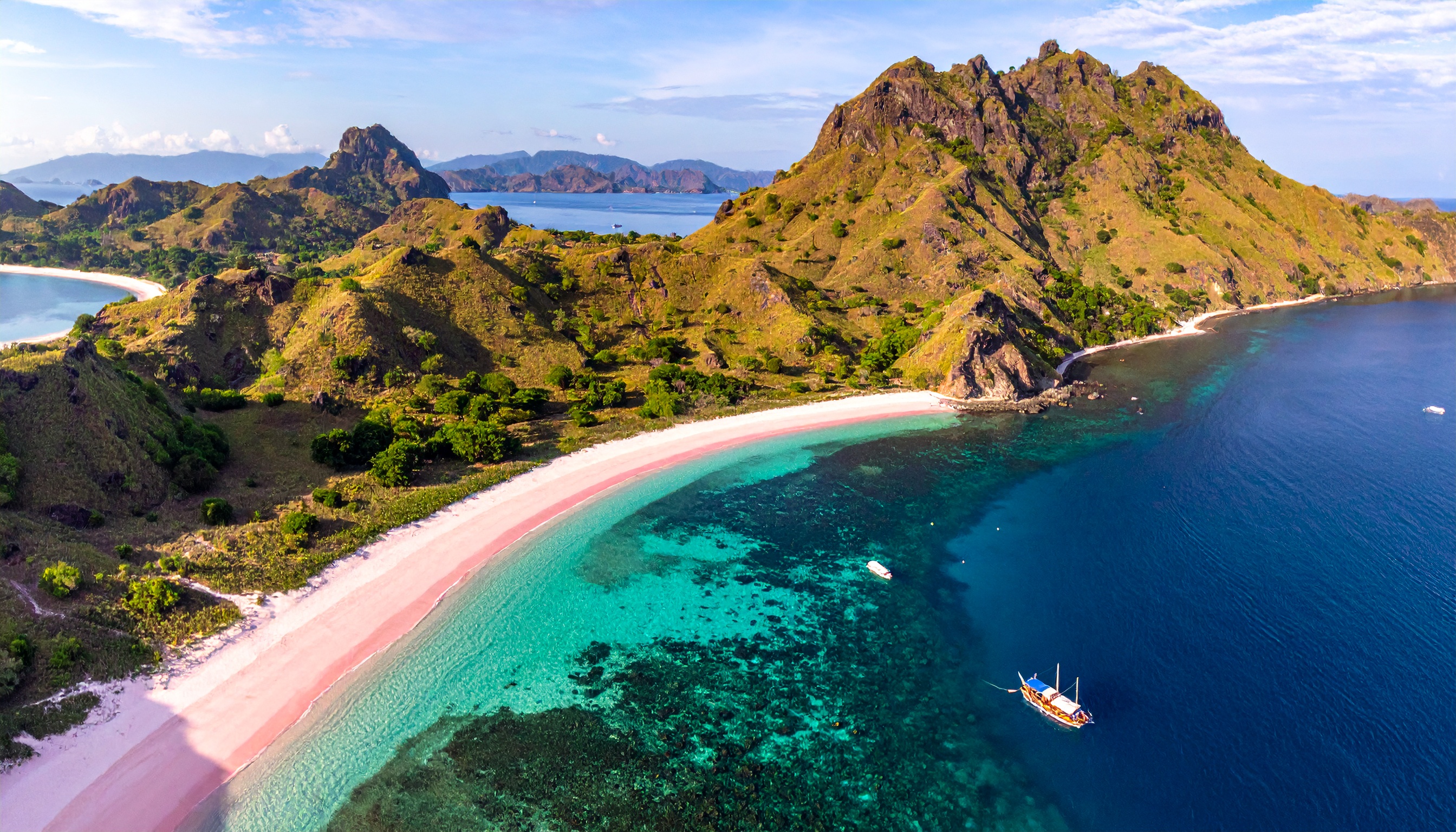 Praia de areia rosada em meio a montanhas e mar azul-turquesa. Ideal para turismo ecológico e relaxamento em ambientes naturais. A paisagem conta com colinas verdes, água cristalina e uma embarcação à deriva. Perfeita para quem busca férias paradisíacas e experiências únicas ao ar livre. Uma escolha excepcional para fotos de viagem e maximizar a tranquilidade.