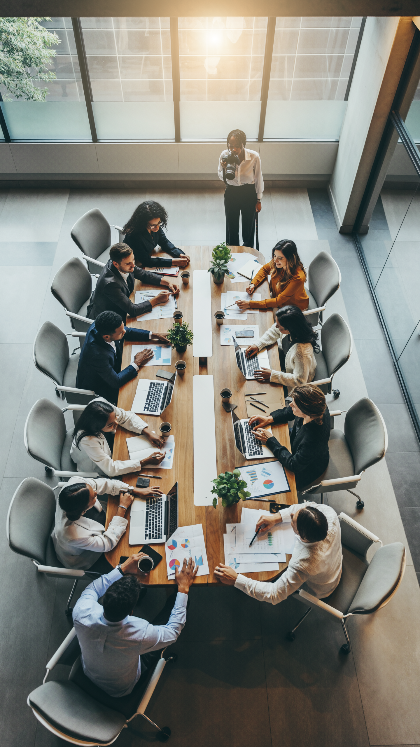 Cena de uma reunião de negócios em uma sala moderna com várias pessoas ao redor de uma mesa retangular, usando laptops e analisando gráficos. A iluminação é natural, vinda de grandes janelas ao fundo, criando uma atmosfera profissional e colaborativa. O ângulo de visão é de cima, proporcionando uma visão clara da interação entre os membros da equipe.