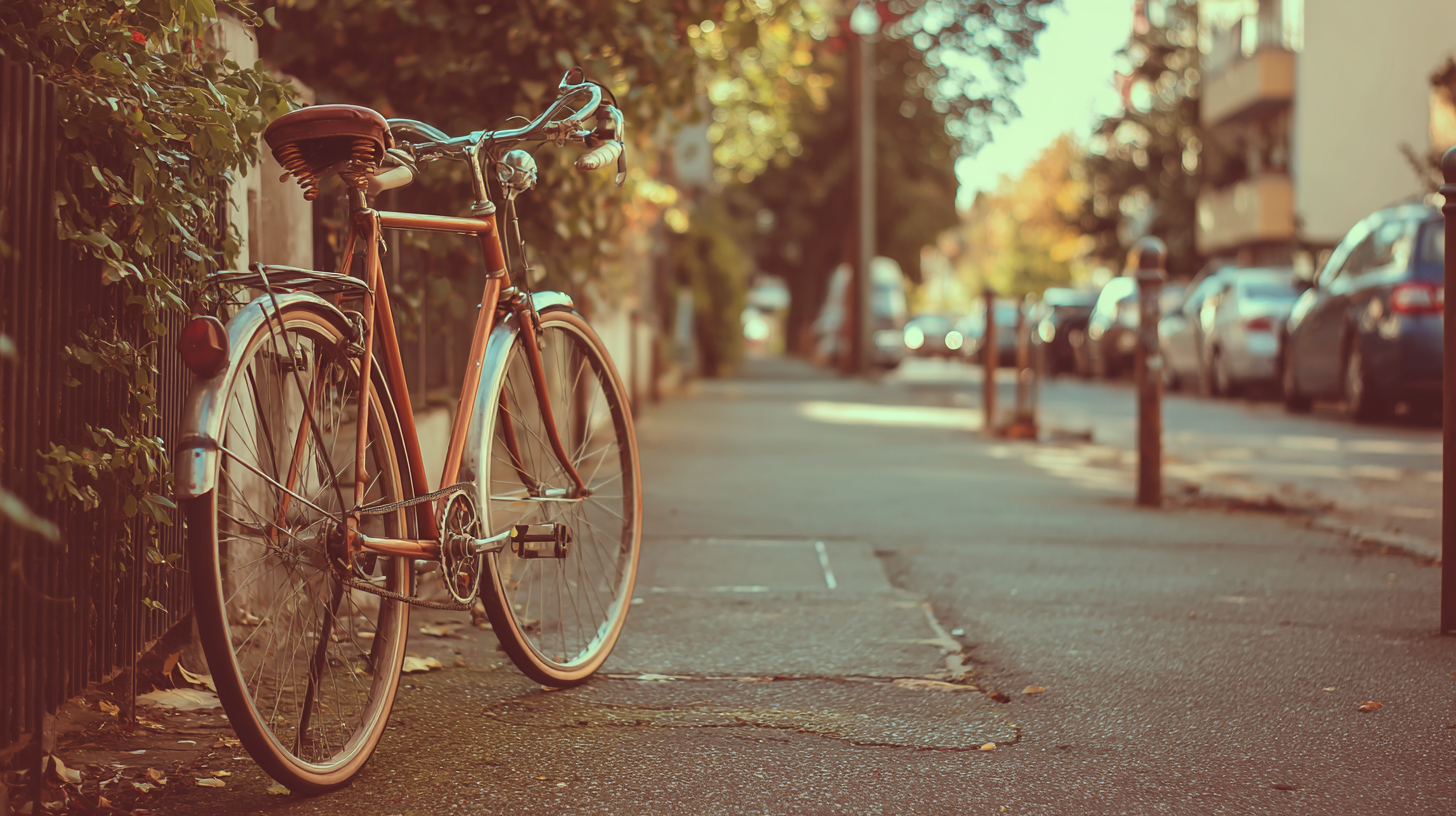 Uma bicicleta vintage laranja estacionada em uma calçada com folhas caídas, rodeada por vegetação e carros ao fundo em uma rua urbana durante o outono.