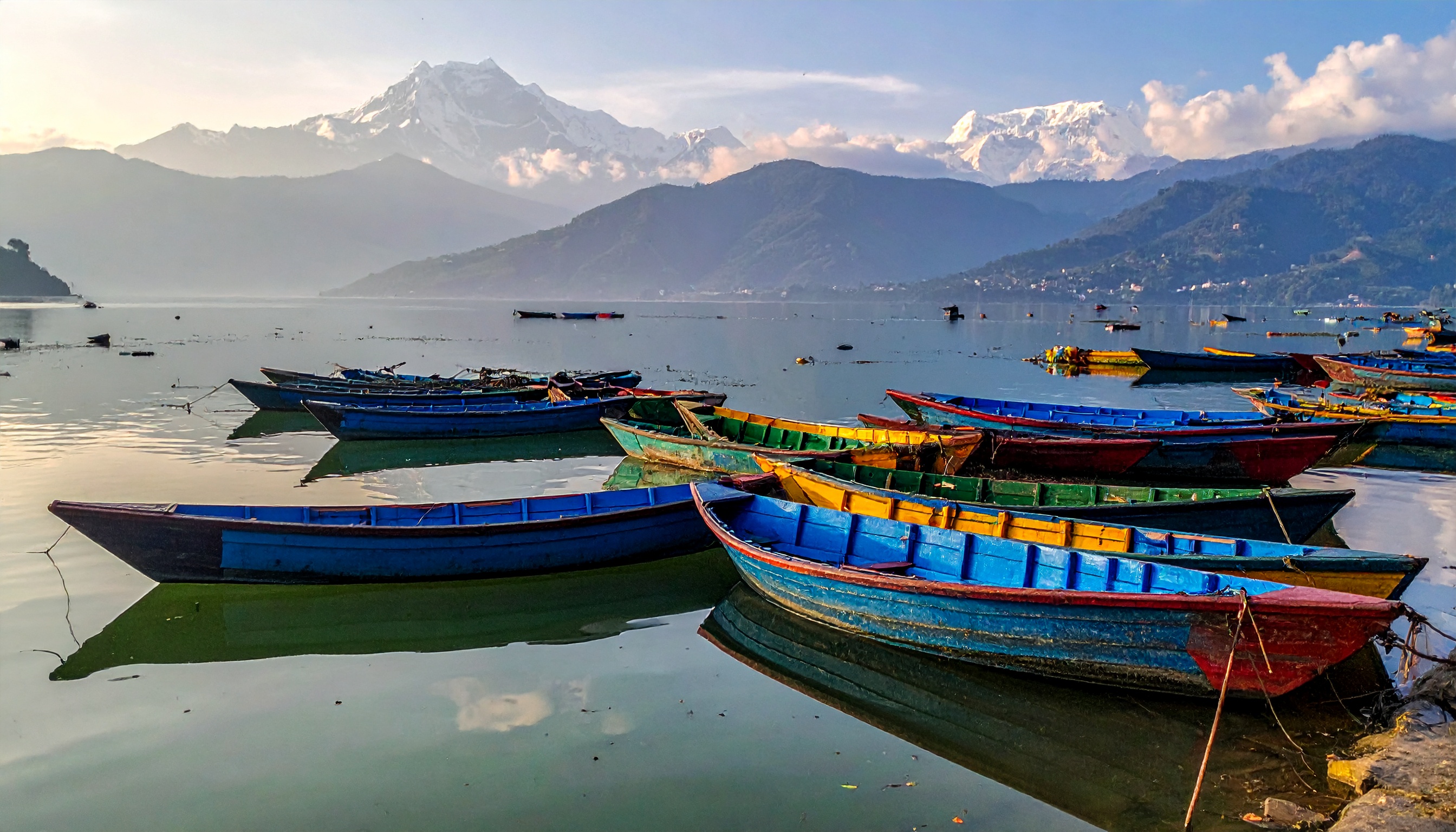 Barcos coloridos flutuam em um lago tranquilo com montanhas ao fundo, criando uma cena pitoresca. Ideal para promoção de turismo em regiões montanhosas ou destinos de ecoturismo. Os barcos em azul e verde contrastam vivamente com a água serena e as montanhas cobertas de neve ao fundo. Perfeito para uso em materiais promocionais de viagens durante o verão ou primavera. A imagem destaca a harmonia entre natureza e atividade humana, proporcionando uma sensação de calma e aventura.