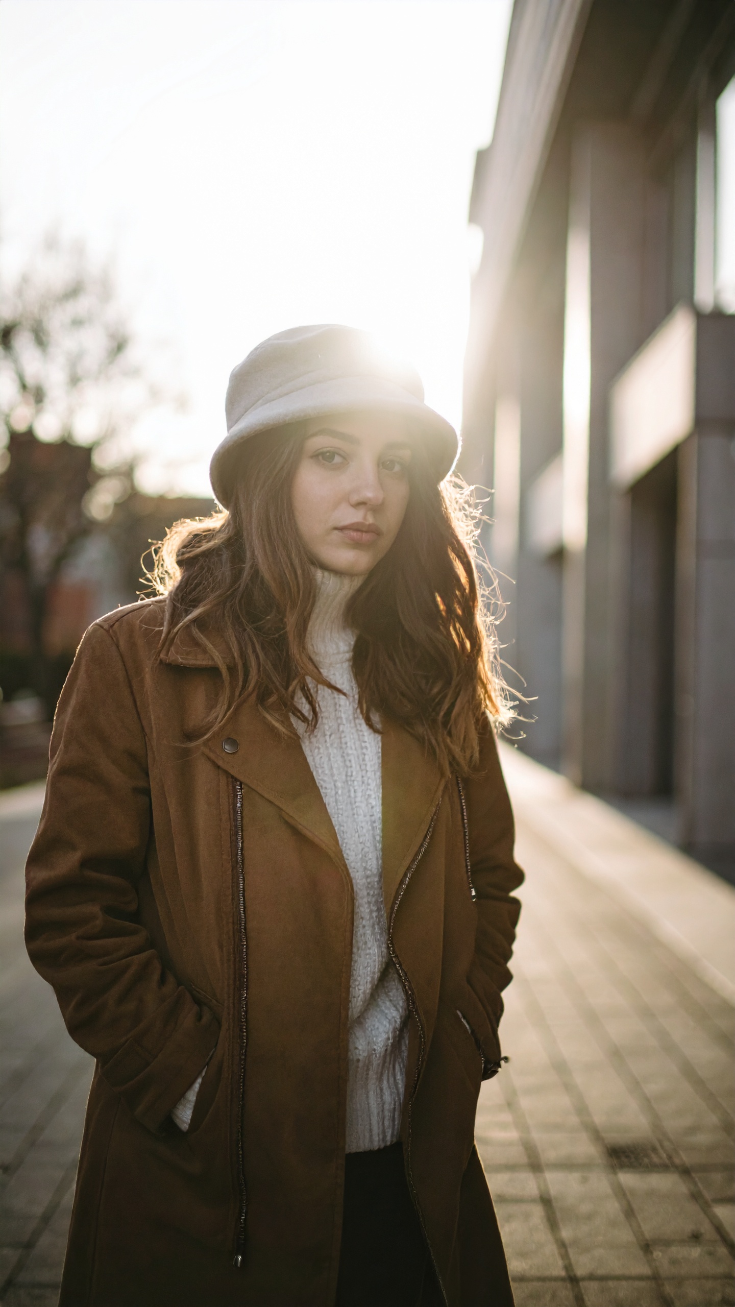 Young woman in a stylish coat and hat stands in urban sunlight