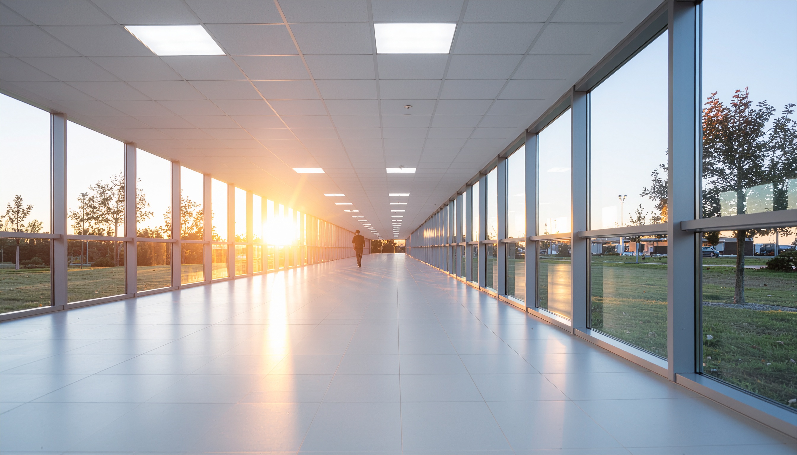 Corredor moderno com janelas de vidro em ambos os lados, captando a luz suave do pôr-do-sol, refletida no piso brilhante. A composição simétrica e perspectiva linear criam uma sensação de profundidade, enquanto uma pessoa caminha ao fundo. A iluminação clara destaca o design minimalista e o ambiente tranquilo.