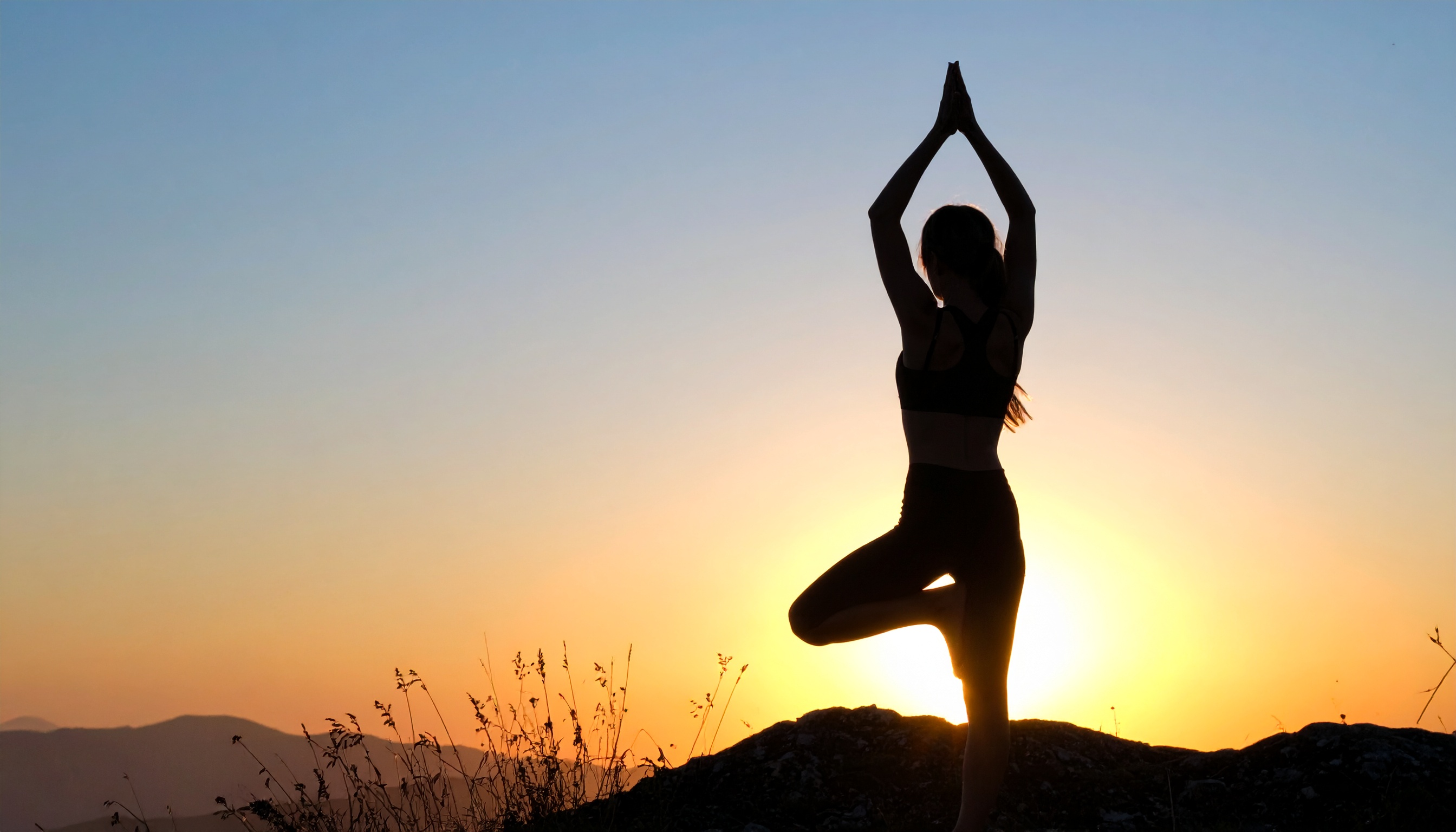 Silhueta de uma pessoa praticando yoga ao pôr do sol, em posição de árvore, sobre uma colina. A cena destaca o contraste dramático entre o céu alaranjado do entardecer e a figura escura, criando uma atmosfera serena e meditativa. A composição é centrada e equilibrada, com foco no equilíbrio da pose contra a paisagem natural ao fundo.