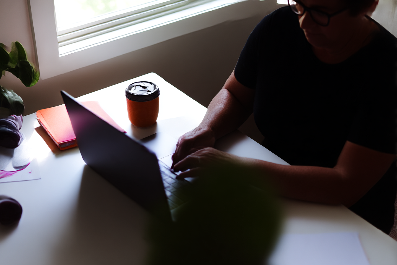 Pessoa trabalhando em um laptop em uma mesa iluminada, com uma caneca laranja e cadernos coloridos ao lado. O ambiente é acolhedor e bem iluminado, ideal para produtividade.