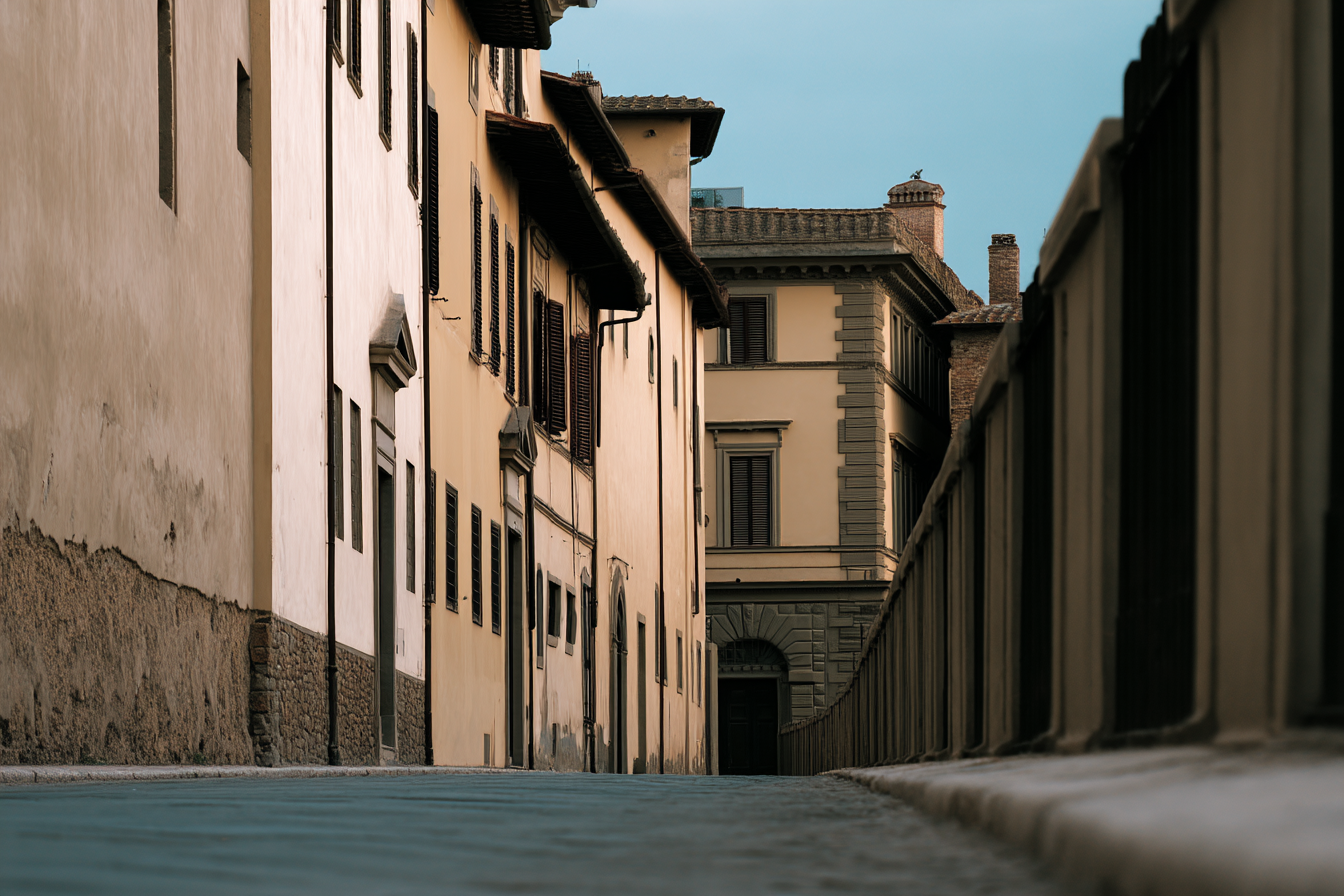 Rua estreita e deserta com edifícios históricos de fachadas clássicas e janelas de madeira, iluminada suavemente pela luz do fim de tarde, criando uma atmosfera tranquila e nostálgica. A perspectiva em ângulo baixo destaca a textura das paredes e o alinhamento arquitetônico. Céu azul claro complementa a composição simétrica e harmônica.