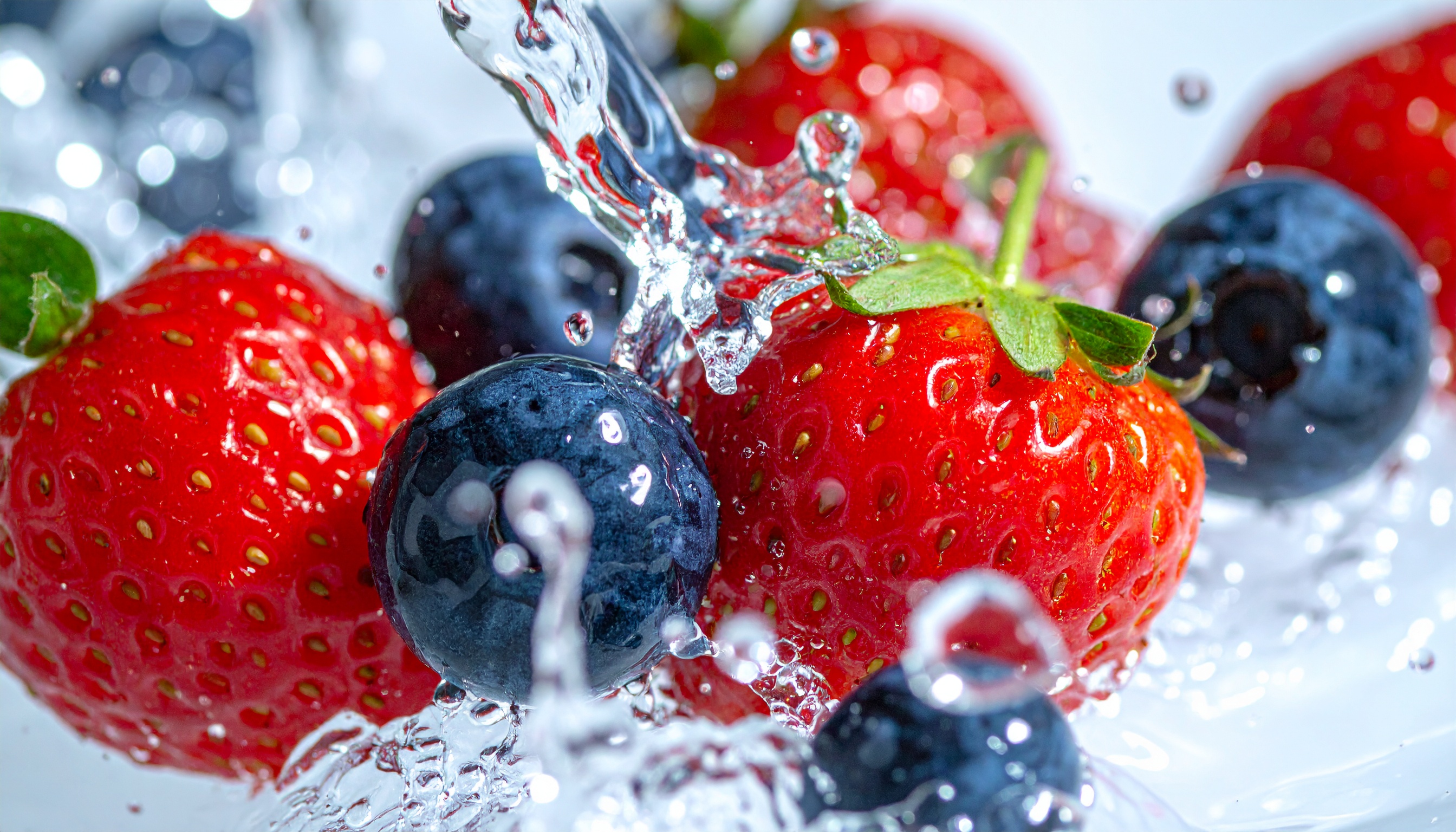 Imagem vívida de morangos e mirtilos frescos sendo salpicados por gotas de água cristalina, destacando suas cores vibrantes e textura suculenta. O close-up captura detalhes como a superfície brilhante das frutas e o dinamismo da água em movimento, proporcionando uma sensação de frescor e vitalidade. A iluminação clara e direta enfatiza o contraste entre o vermelho dos morangos e o azul dos mirtilos, criando uma composição atraente e refrescante.
