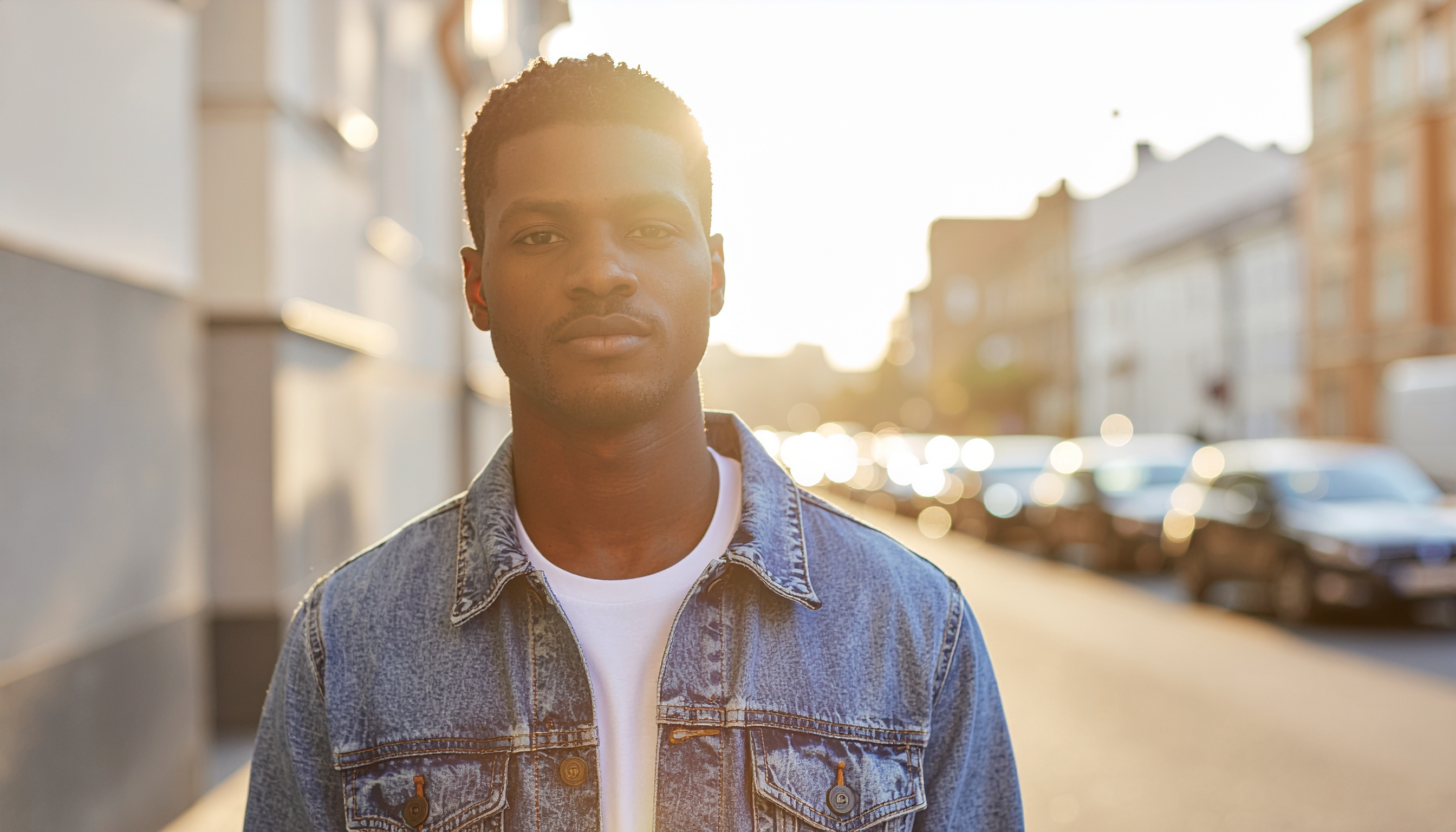 Retrato de um homem em uma rua urbana ao entardecer, usando jaqueta jeans. A iluminação suave do pôr do sol cria um efeito de silhueta, destacando o rosto. O fundo desfocado sugere uma cidade em movimento, com carros e edifícios, criando uma atmosfera tranquila e contemplativa.