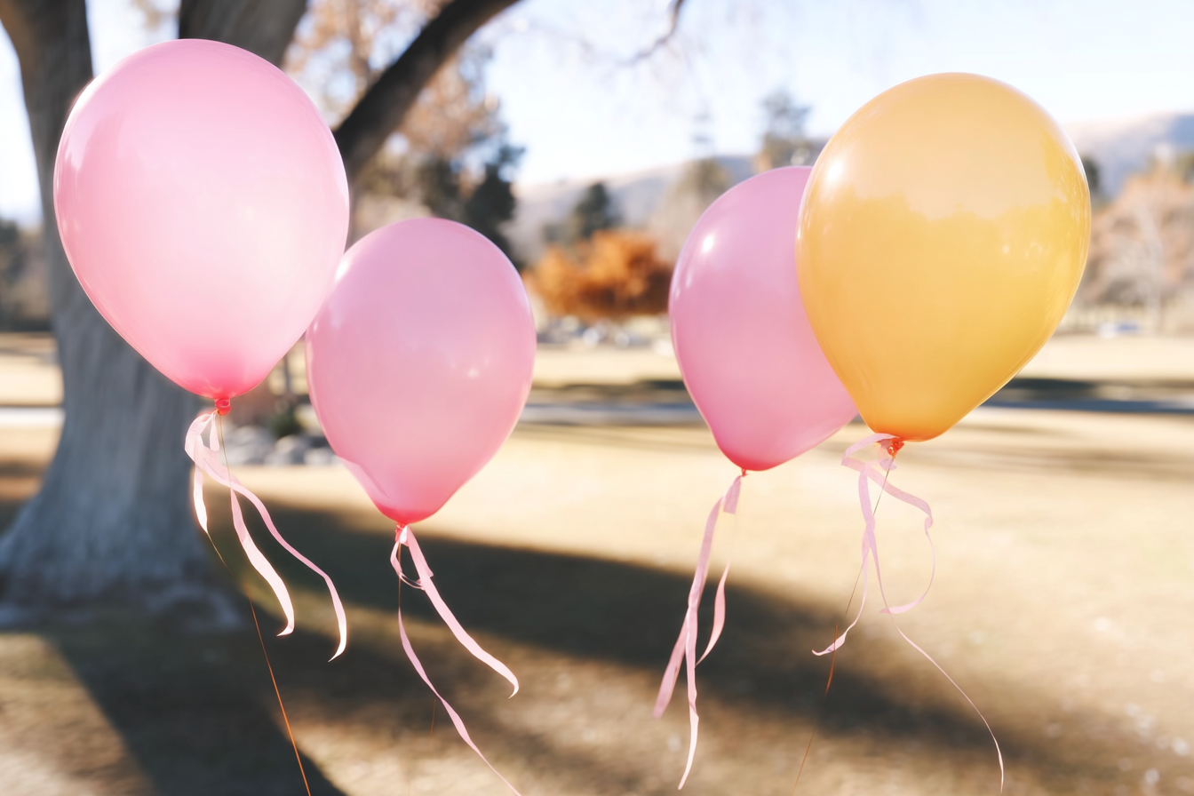 Quatro balões coloridos em um parque, incluindo balões rosa e um balão amarelo, flutuando suavemente com fitas penduradas, em um ambiente ensolarado com árvores ao fundo.