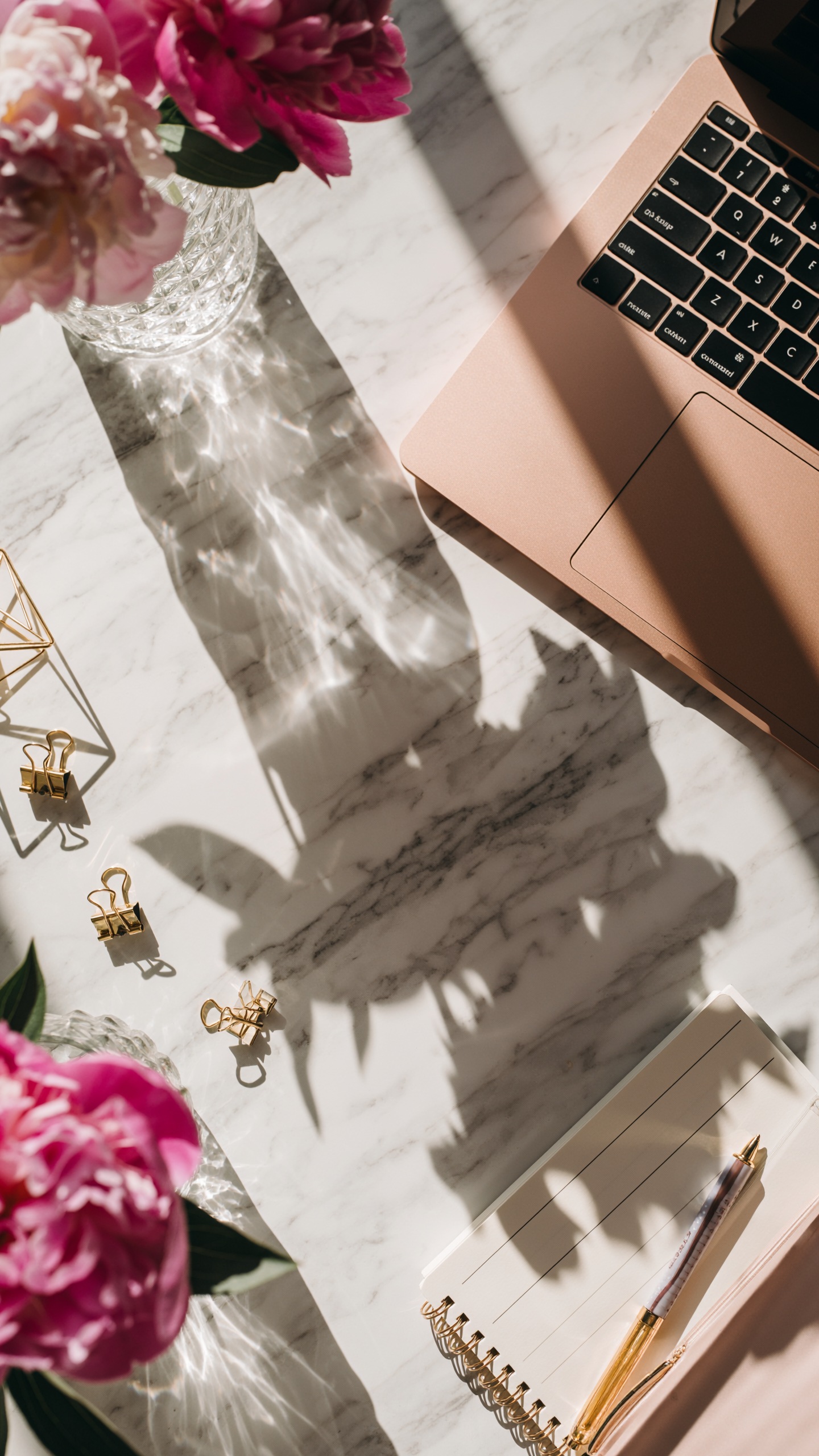 Cena de uma mesa elegante com laptop em tom rosé, flores vibrantes em vaso de cristal, clipes dourados, caderno espiral e caneta, sob luz natural suave que projeta sombras artísticas sobre a superfície de mármore branco.