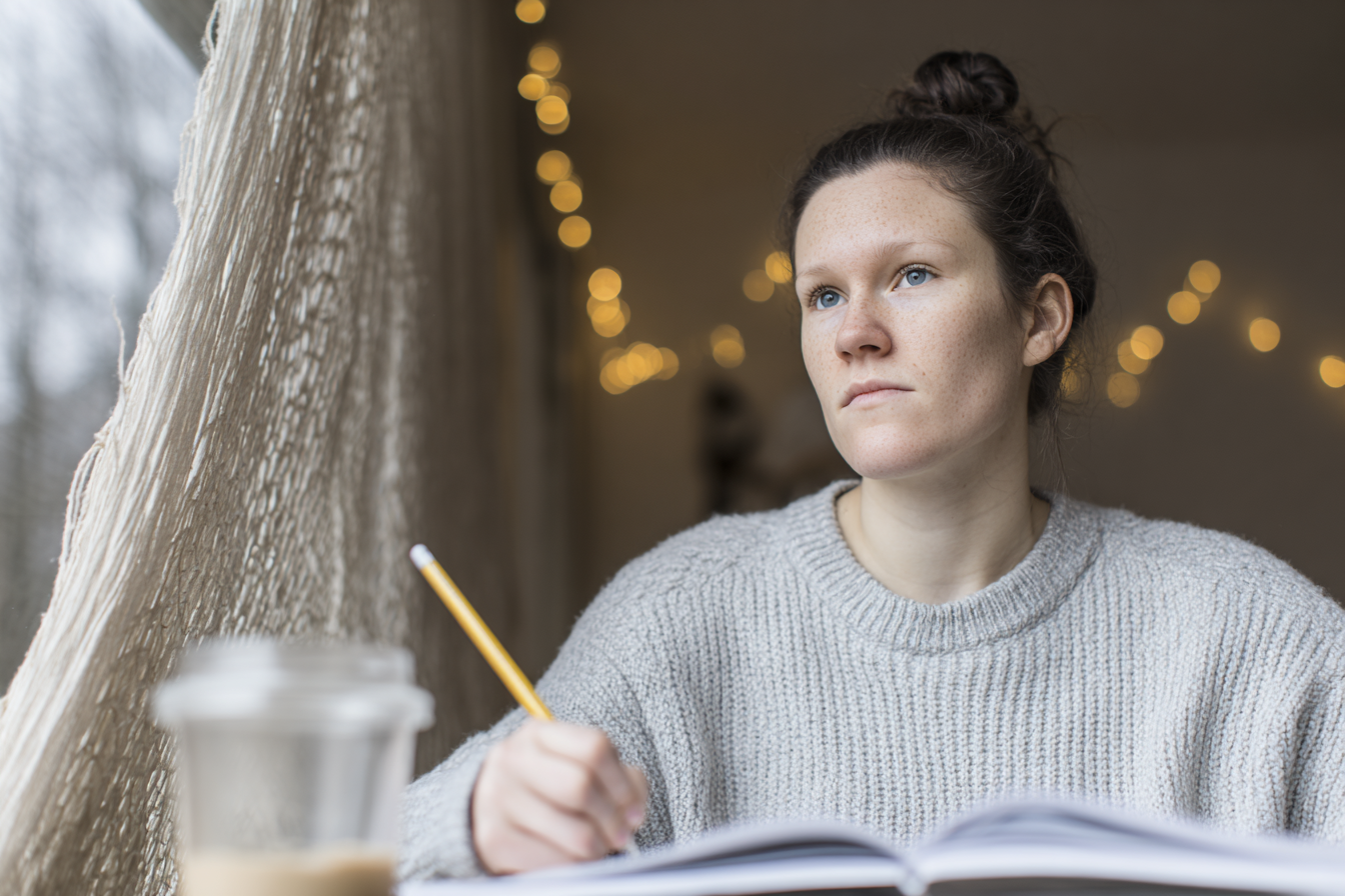 Mulher pensativa escreve em caderno enquanto olha pela janela com luzes de natal ao fundo, criando uma atmosfera contemplativa e acolhedora.