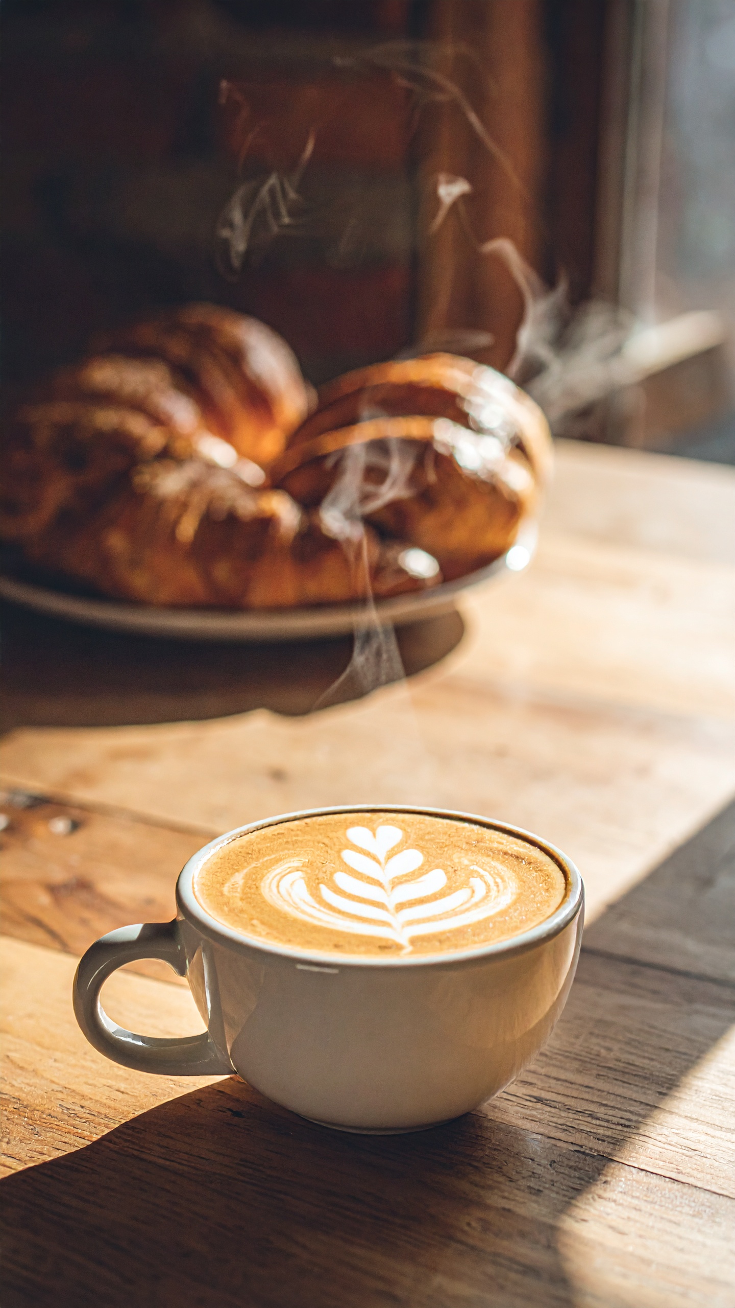 Café latte com arte em espuma em mesa de madeira aquecida pelo sol. Ideal para momentos de relaxamento ou encontros em cafeterias. A xícara branca contrasta com o padrão intrincado da espuma. Perfeito para um brunch ou café da manhã acolhedor. A combinação de luz natural e textura do latte cria um ambiente convidativo.