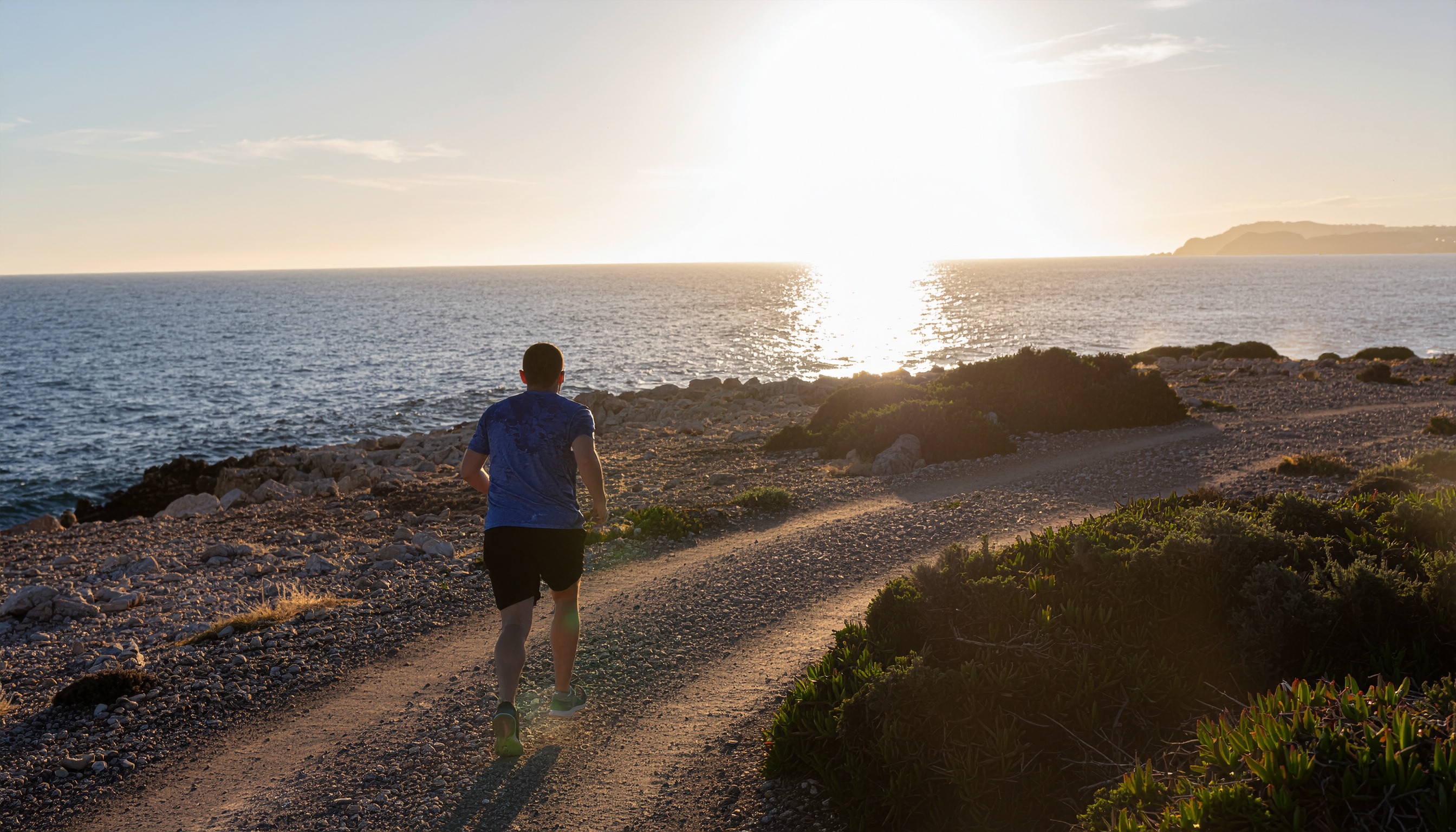 Homem correndo em uma trilha costeira ao pôr do sol, com o oceano à esquerda refletindo a luz dourada. A cena tem uma atmosfera serena e energizante, com iluminação natural suave e composição harmoniosa. Vegetação rasteira e pedras compõem o cenário natural, enquanto o ângulo baixo destaca a textura do caminho.