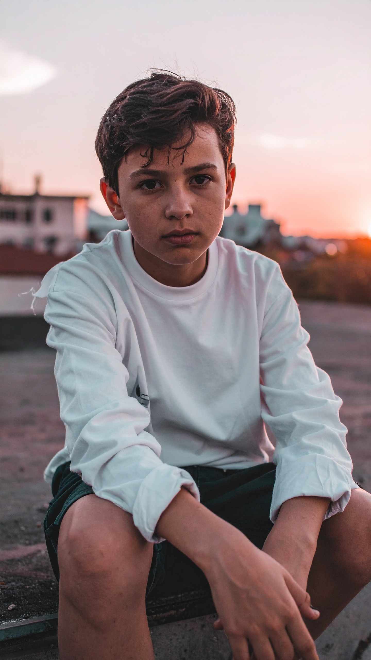 Teenage boy in a casual white shirt sits thoughtfully on a rooftop at sunset
