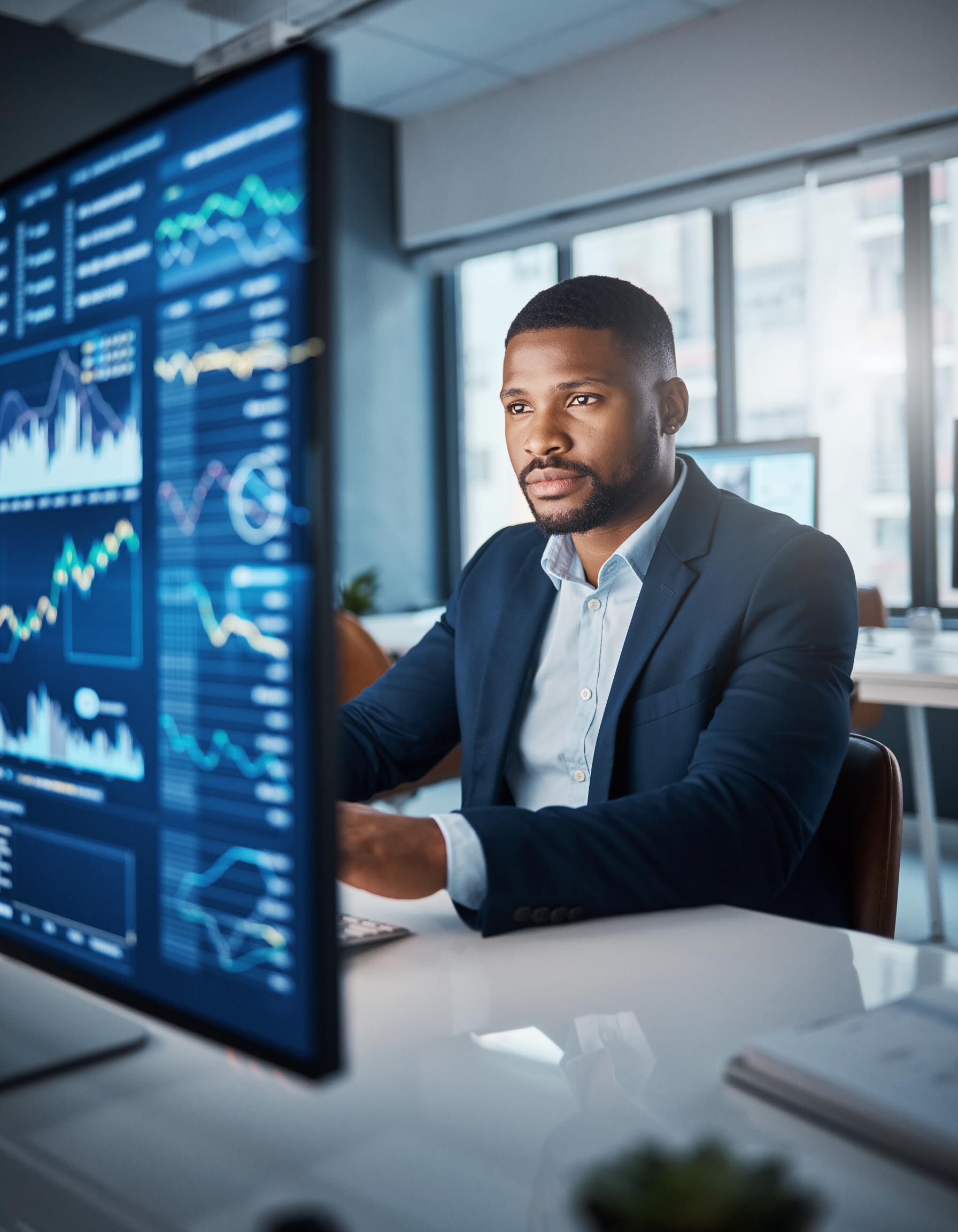 Homem em traje formal analisa gráficos financeiros em tela de computador em escritório moderno, transmitindo foco e profissionalismo em ambiente corporativo avançado.