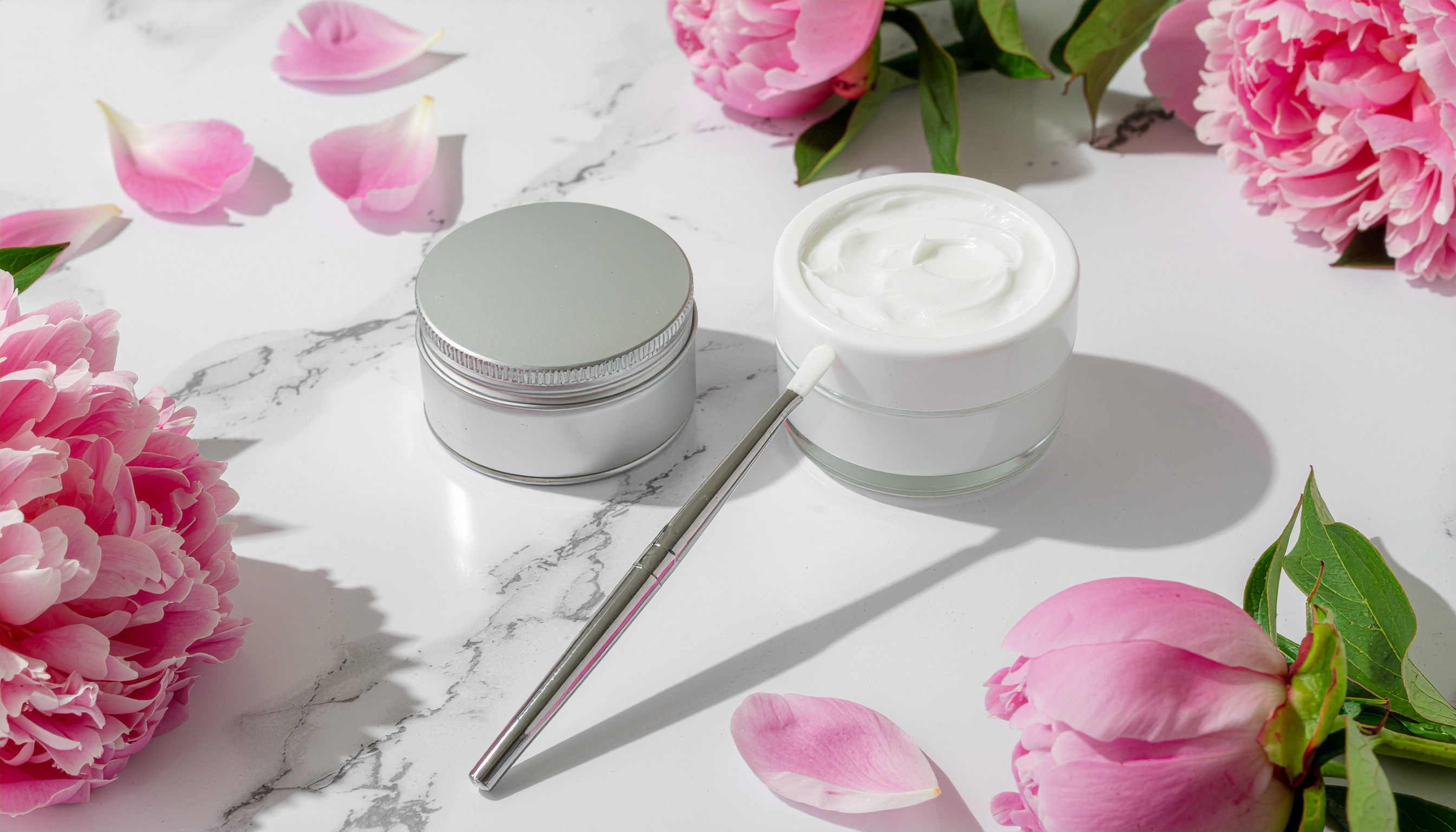 Creme em pote branco ao lado de lata metálica sobre superfície de mármore com flores de peônia rosa e pétalas, em um ambiente bem iluminado que destaca a textura cremosa e os detalhes florais, criando uma composição elegante e suave.