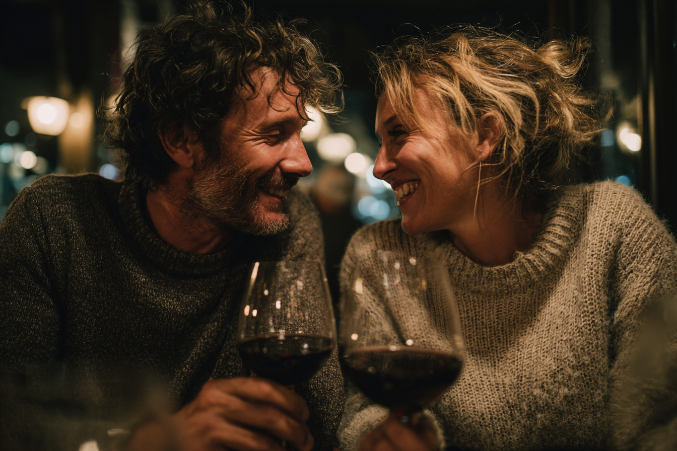 A imagem retrata um momento caloroso entre um casal em um ambiente aconchegante. As cores predominantes incluem tons quentes e terrosos, com um foco especial em marrons, cinzas e os reflexos do vinho tinto. As formas são suaves e arredondadas, com o destaque para os rostos sorridentes dos indivíduos e as taças de vinho. A composição é íntima, com os dois personagens centralizados em um ambiente que sugere um restaurante acolhedor. Texturas como a lã dos suéteres e a transparência do vidro das taças são notáveis, criando um contraste agradável. O padrão de luz suave ao fundo contribui para a atmosfera romântica e relaxante.