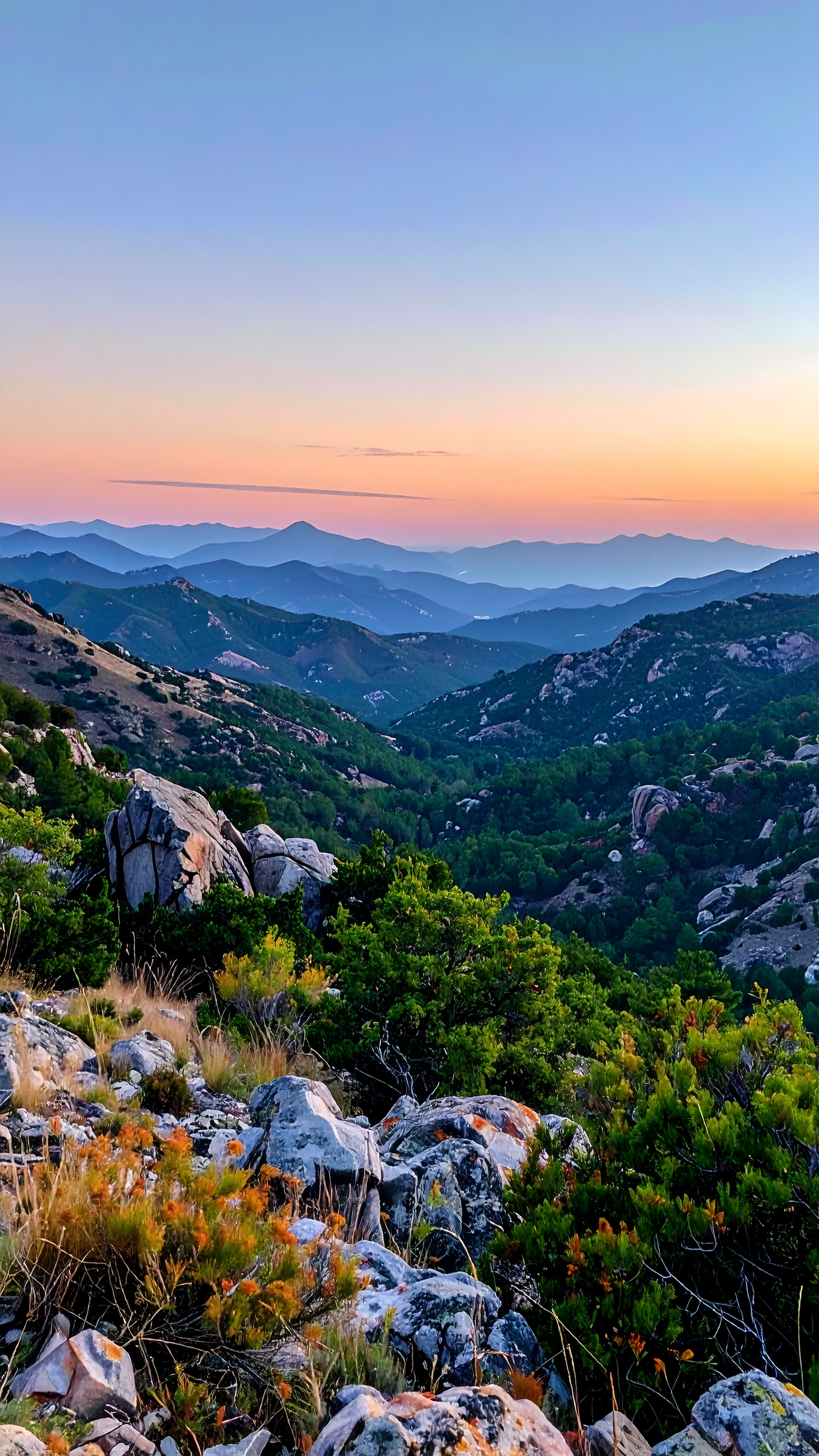 Paisagem montanhosa com vegetação esparsa e rochas em primeiro plano, estendendo-se até o horizonte com várias camadas de montanhas sob um céu colorido pelo pôr do sol, criando uma atmosfera serena e majestosa.