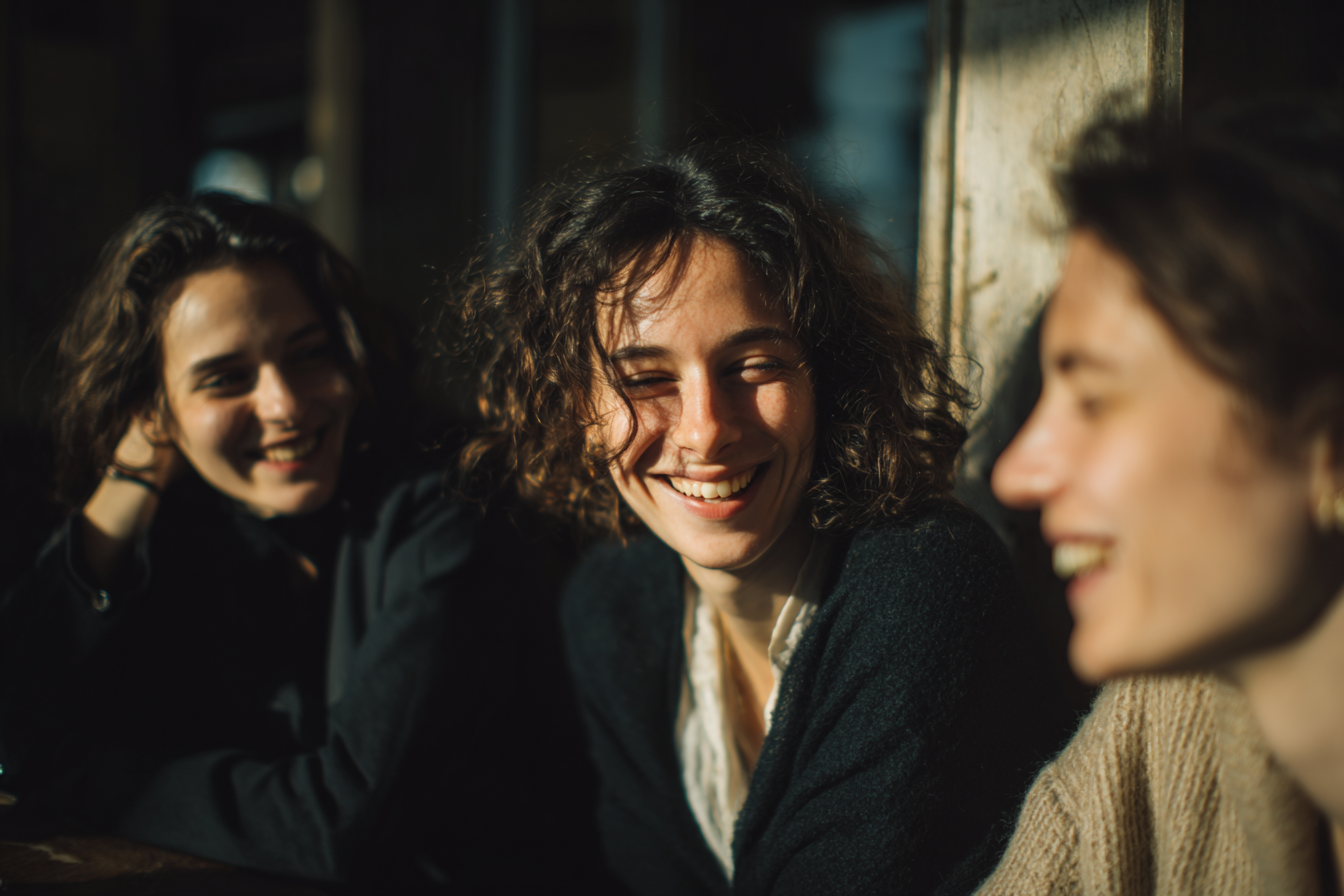 Três mulheres sorrindo e se divertindo em um ambiente iluminado, com luz suave refletindo em seus rostos e cabelo cacheado ao sol. A imagem captura um momento de alegria e amizade, destacando expressões felizes e uma atmosfera acolhedora.