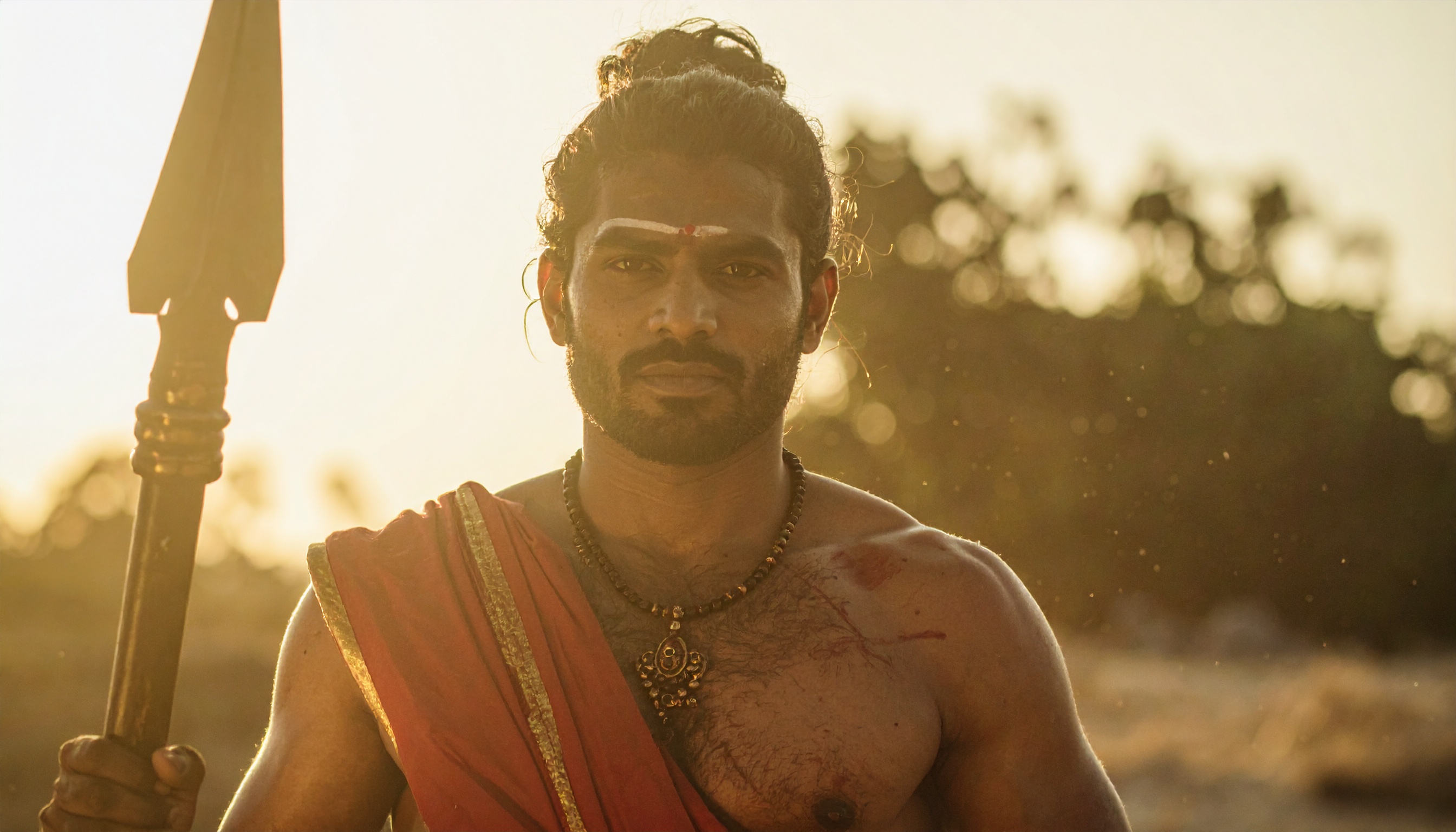 A person in traditional attire holds a spear under a warm, golden sunset