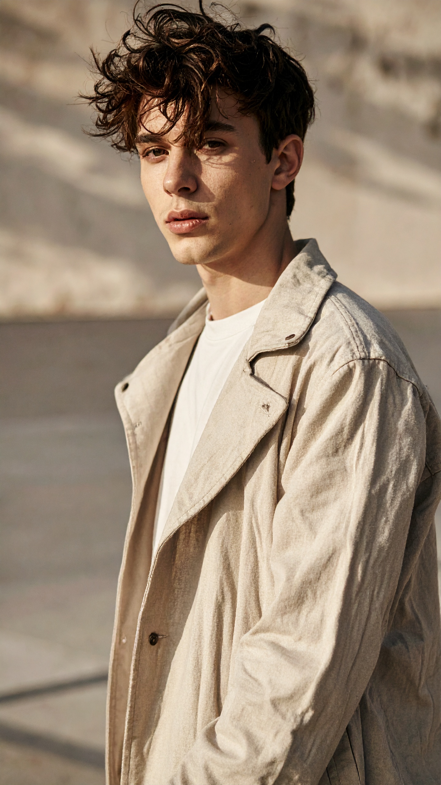 Jovem vestindo casaco bege em ambiente externo ensolarado. Ideal para quem busca estilo casual e despojado, mantendo elegância. O cabelo desarrumado e a luz suave criam uma atmosfera natural e autêntica. Perfeito para ocasiões informais ou ensaios fotográficos ao ar livre. O visual ressalta a simplicidade sofisticada do look moderno.