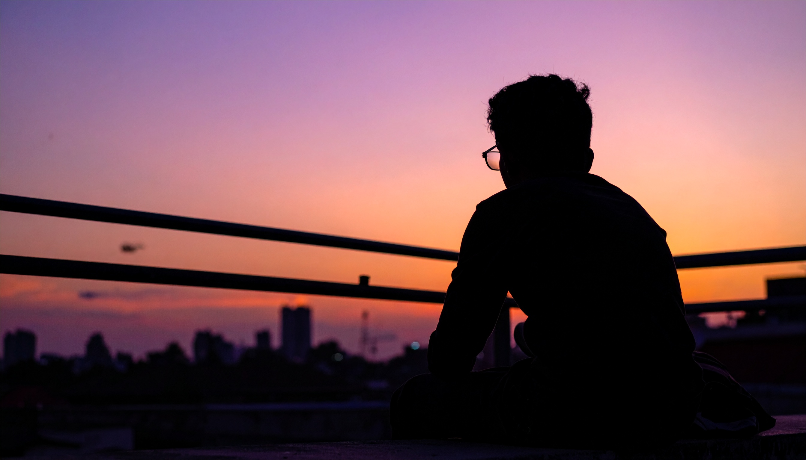 A person sits on a rooftop at sunset, gazing at a colorful sky