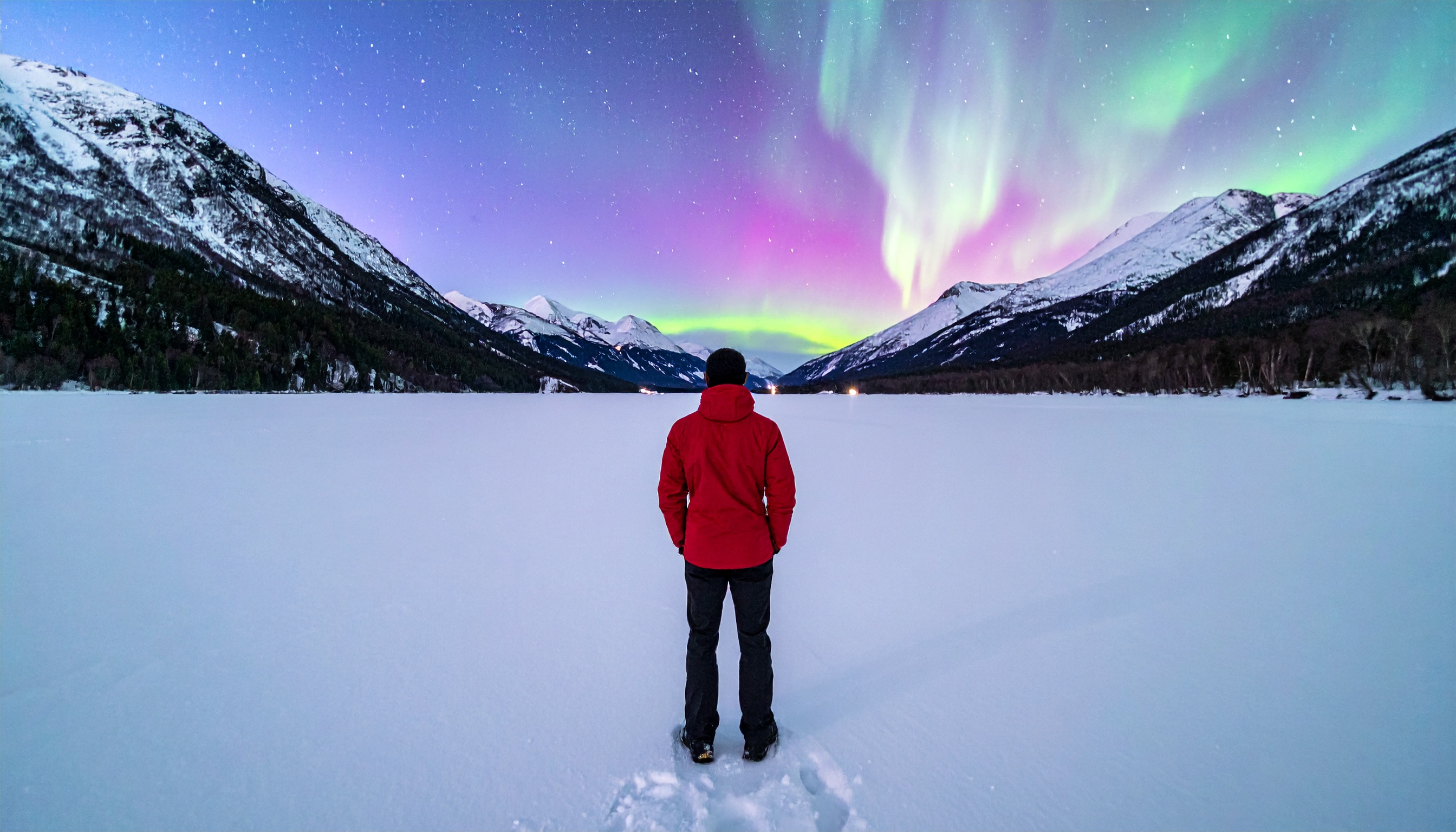 A person in a red jacket stands on a snowy landscape under a vibrant aurora borealis