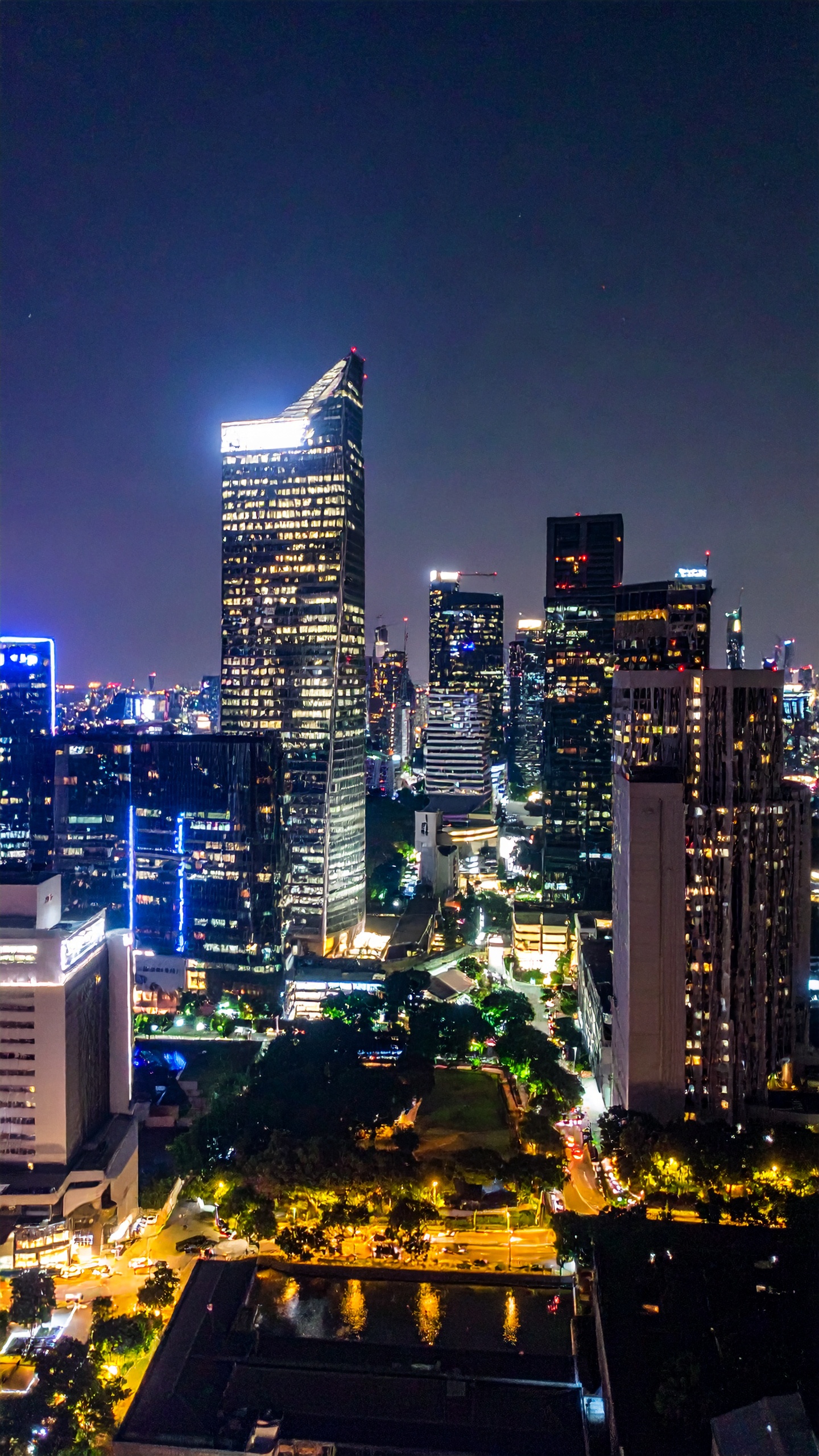 Vista aérea noturna de uma metrópole com arranha-céus iluminados. A imagem captura a energia e a modernidade de uma cidade grande, ideal para campanhas que promovem negócios e tecnologia. Os edifícios altos e brilhantes destacam-se contra o céu escuro, criando um contraste impressionante. Essa cena é perfeita para ilustrar conteúdos sobre vida urbana e inovação. A perspectiva eleva a sensação de dinamismo e progresso, comum em centros urbanos.