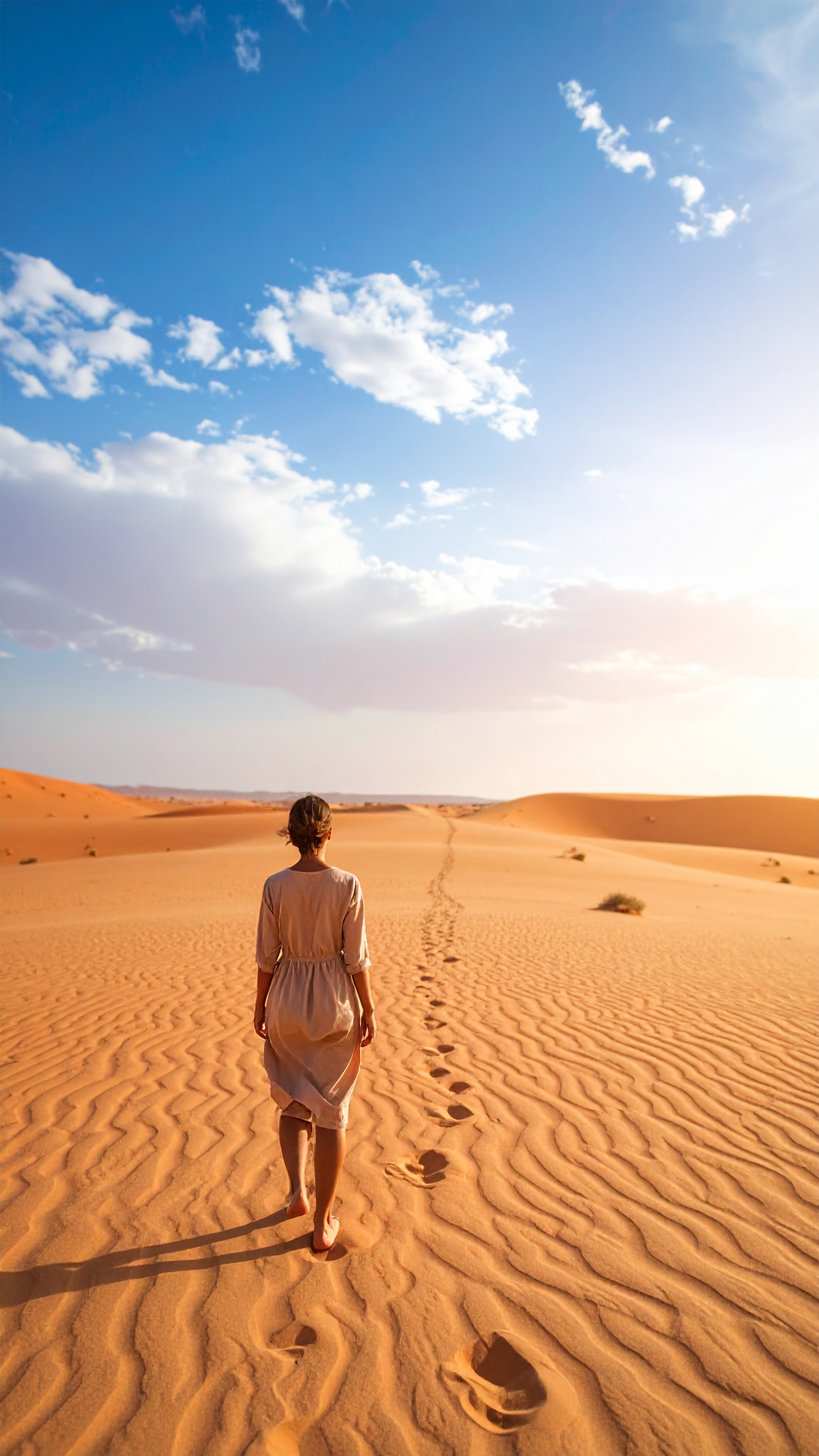 A lone figure walks across vast desert sands under a bright blue sky