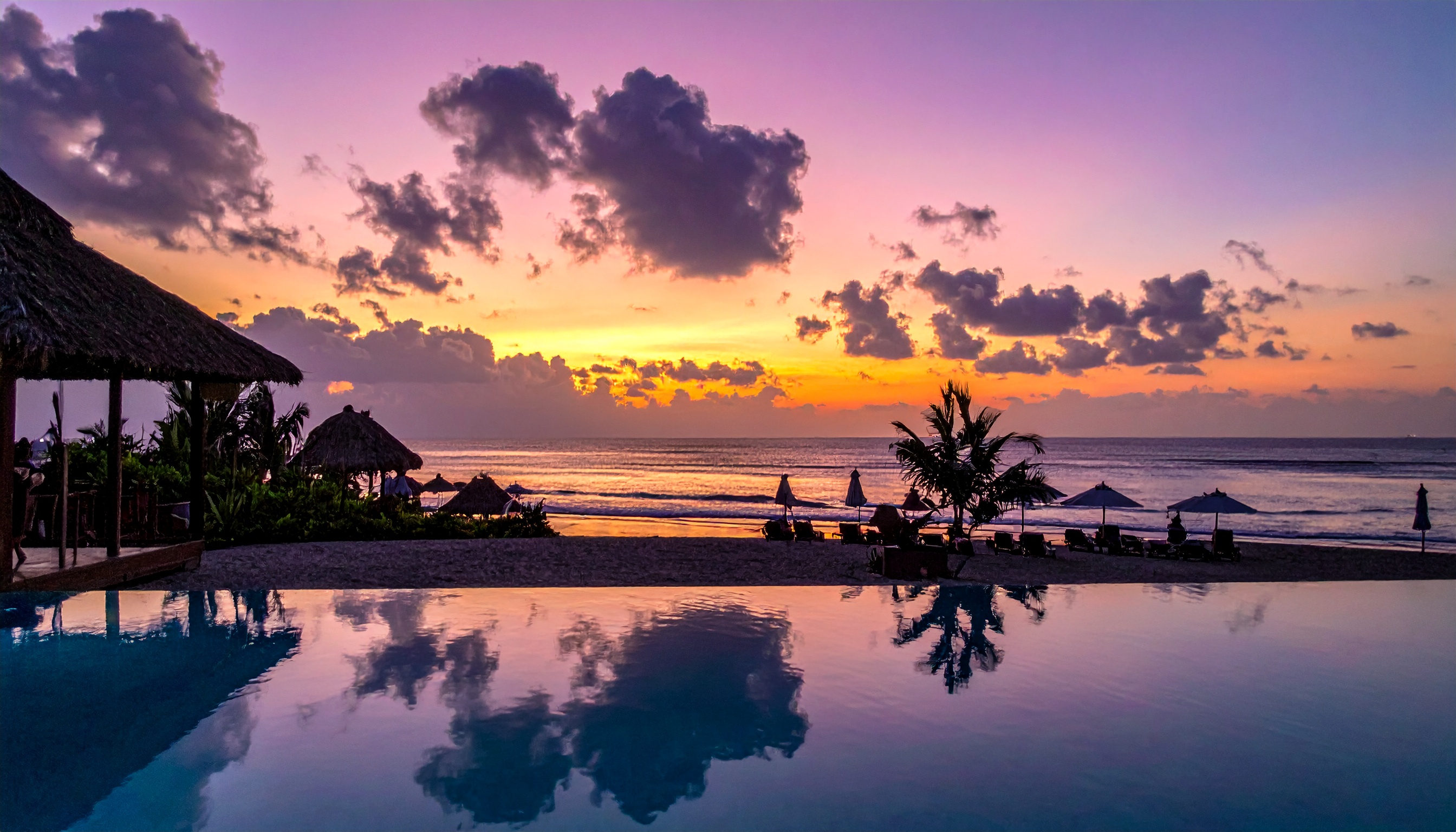 Pôr-do-sol vibrante sobre uma praia serena com ondas suaves e céu colorido. Ideal para quem busca escapar da rotina e relaxar em um cenário tropical. As cores intensas do céu se refletem na água calma da piscina, criando um ambiente de paz e beleza. Perfeito para casais em lua de mel ou viajantes em busca de inspiração. A harmonia entre terra, mar e céu oferece um espetáculo visual único.