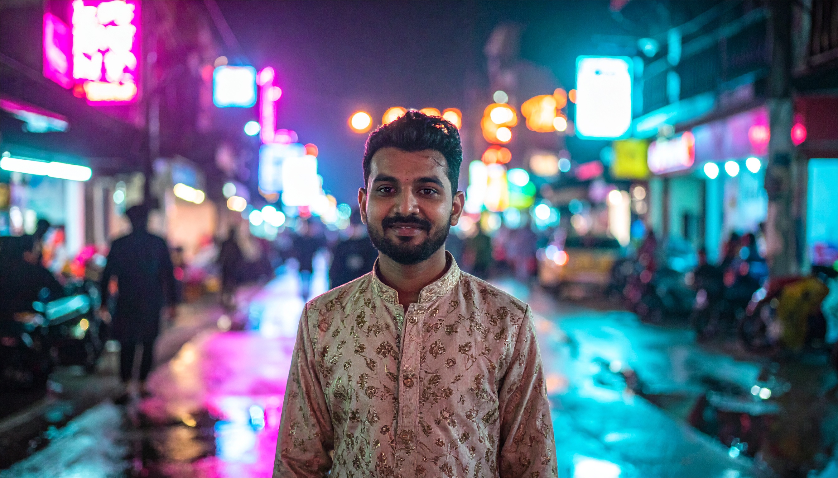 A man wearing an elegant, embroidered shirt stands in a vibrant city street at night