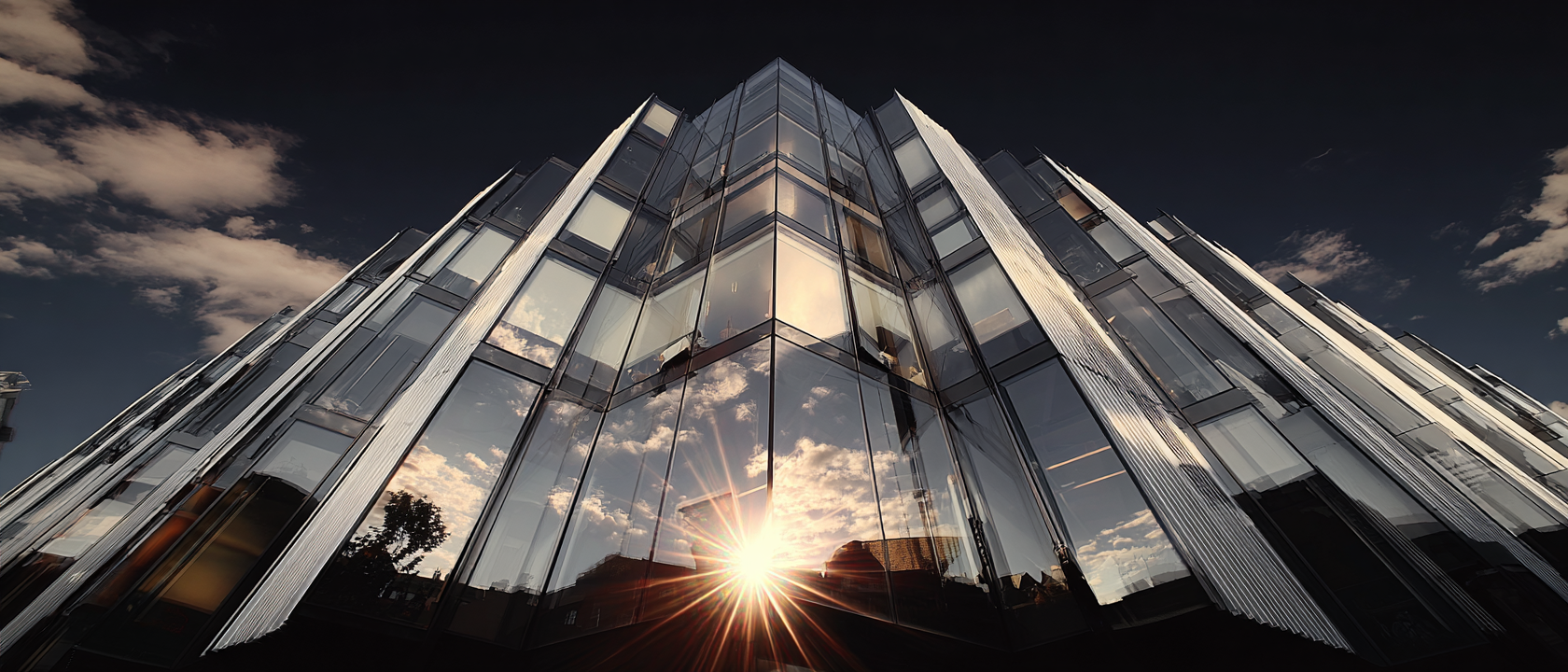Estrutura arquitetônica moderna em vidro refletindo o céu azul e nuvens, iluminada dramaticamente pela luz do sol poente. A composição em ângulo baixo destaca as linhas angulares e o design futurista do edifício, criando um contraste visual impressionante entre a transparência do vidro e a intensidade da luz solar.