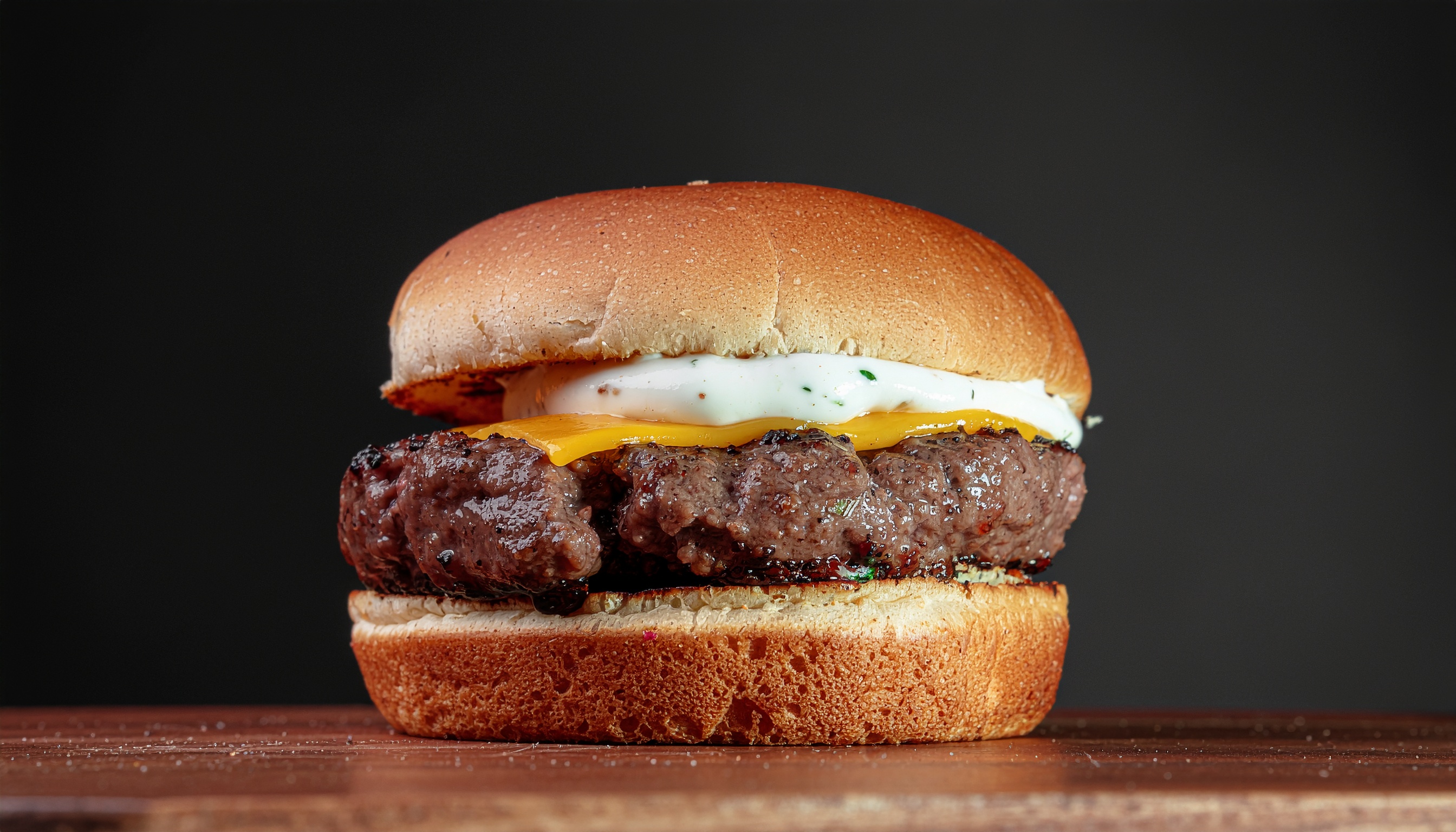 Hambúrguer suculento com queijo derretido e molho cremoso entre pão dourado, sobre superfície de madeira. Iluminação suave destaca textura e cores vibrantes, criando atmosfera apetitosa e convidativa.