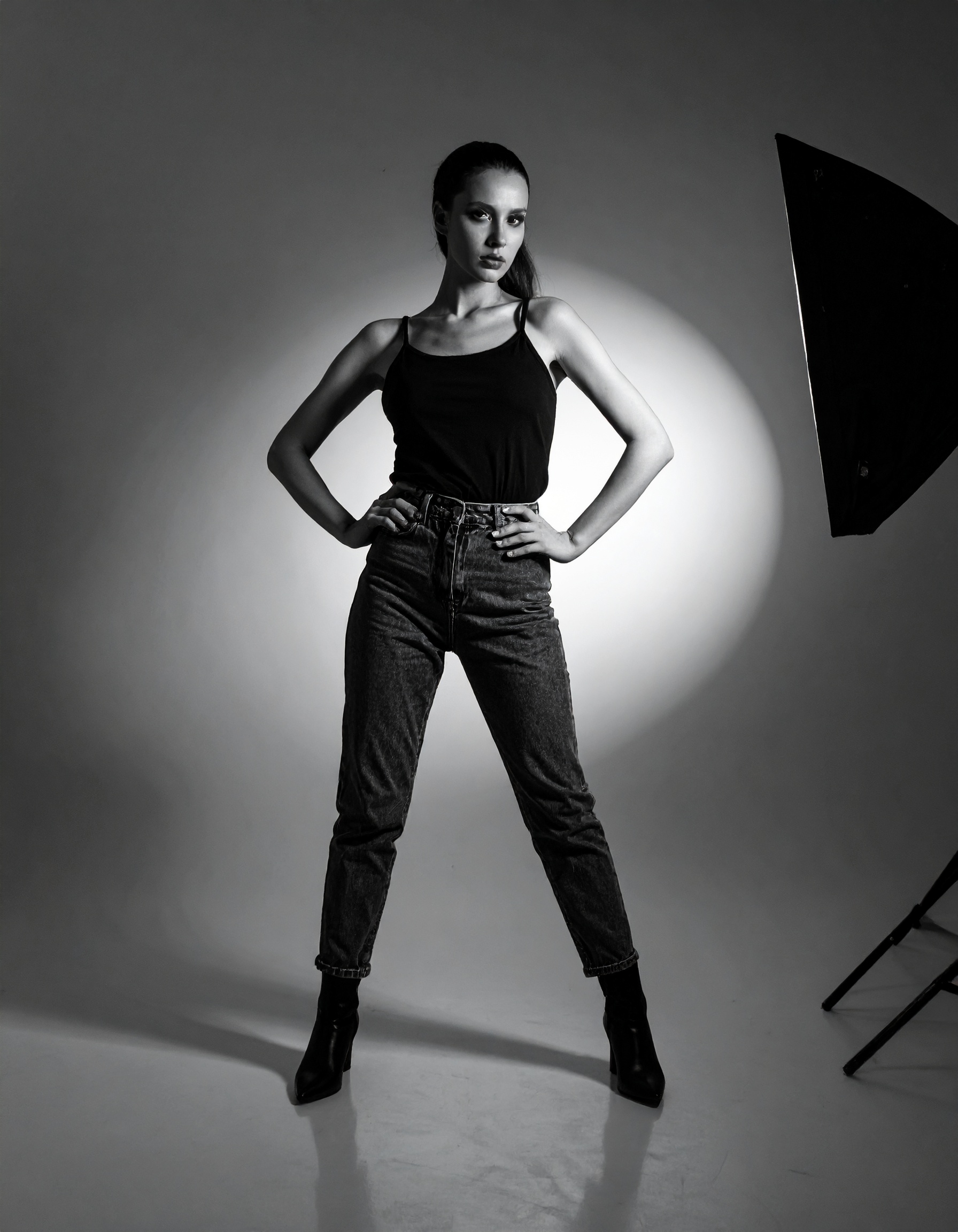 A confident woman poses in a black tank top and jeans against a studio background