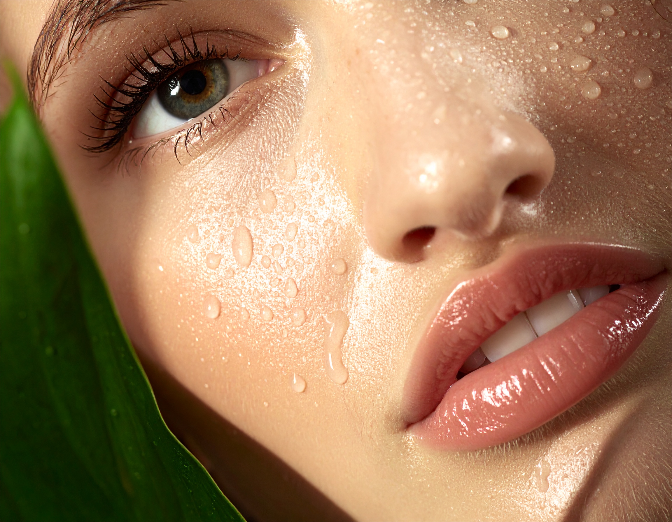 Close-up de rosto feminino com gotículas d'água destacando pele brilhante e olhos expressivos, folha verde ao lado, composição minimalista e refrescante, iluminação suave e foco nítido.