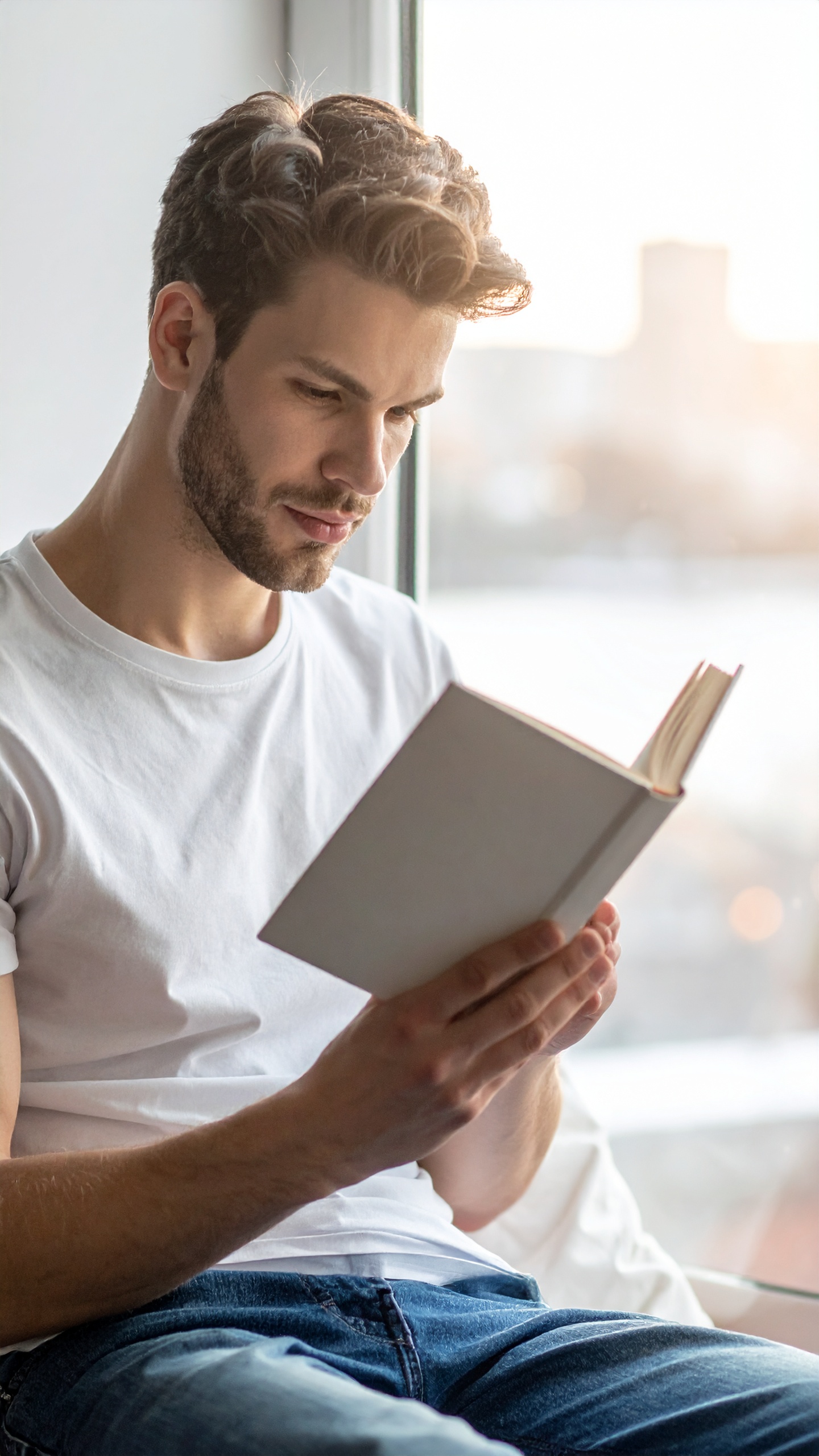 A man sits by a window, engrossed in reading a book