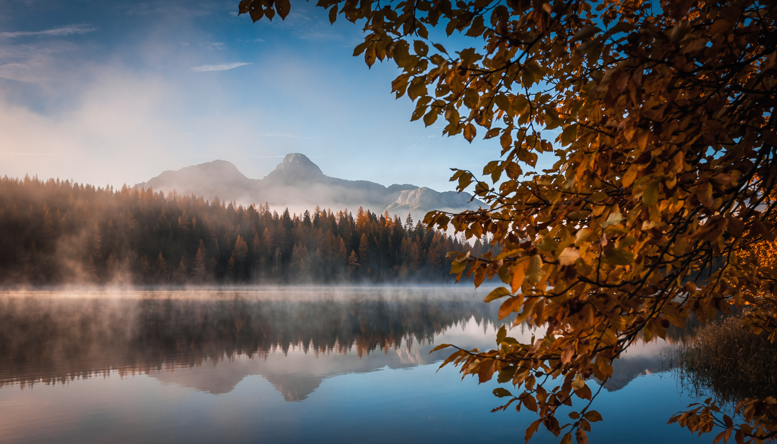 Paisagem serena de outono com folhas douradas em primeiro plano, refletindo em um lago tranquilo cercado por montanhas enevoadas sob um céu azul claro.