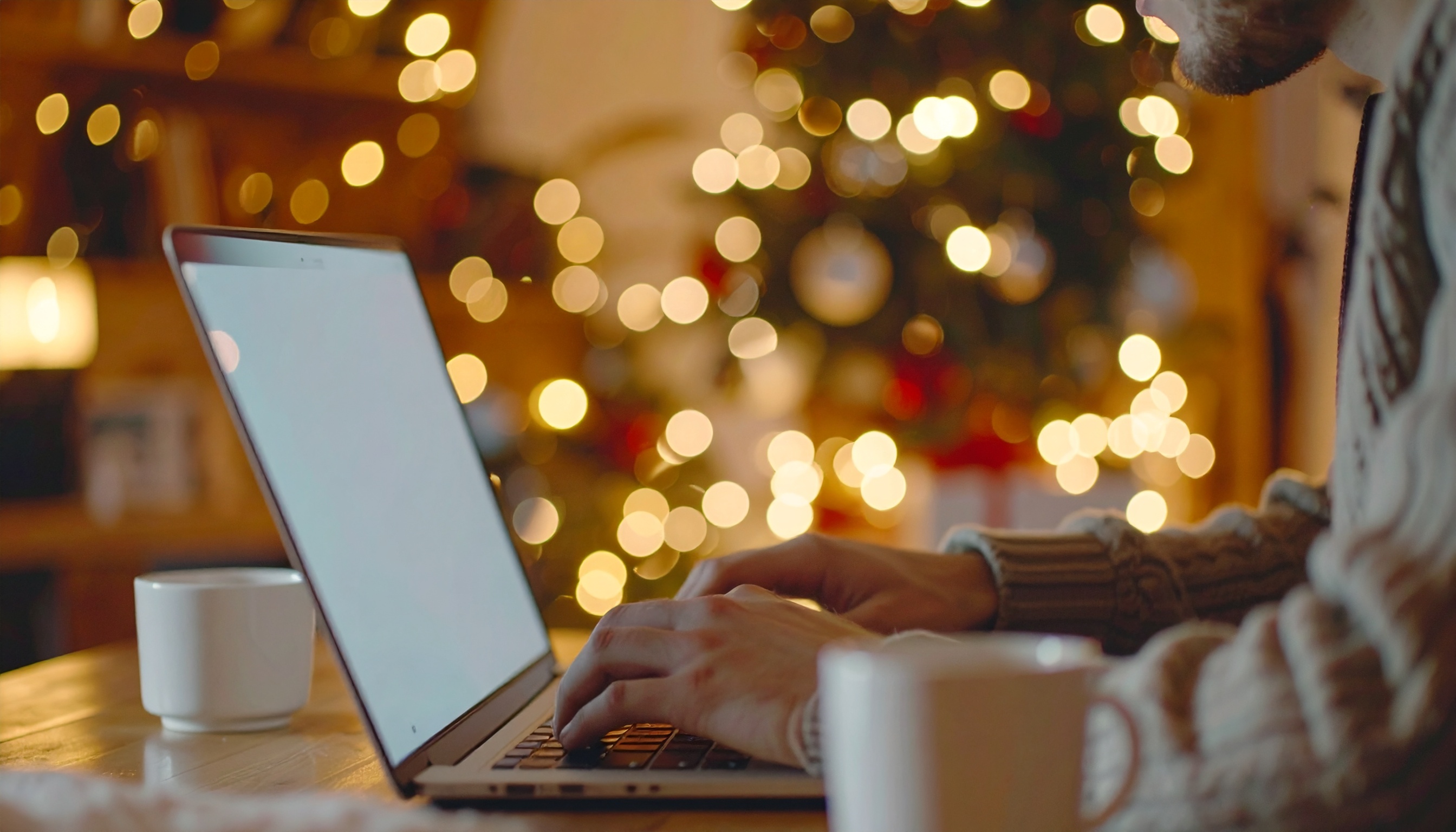 Um ambiente aconchegante com foco em uma pessoa usando laptop em um espaço decorado para o Natal. As luzes de Natal ao fundo criam um efeito bokeh suave, enquanto uma xícara de café está visível sobre a mesa, adicionando um toque de conforto e calor à cena. A iluminação é suave e quente, proporcionando uma atmosfera festiva e tranquila.