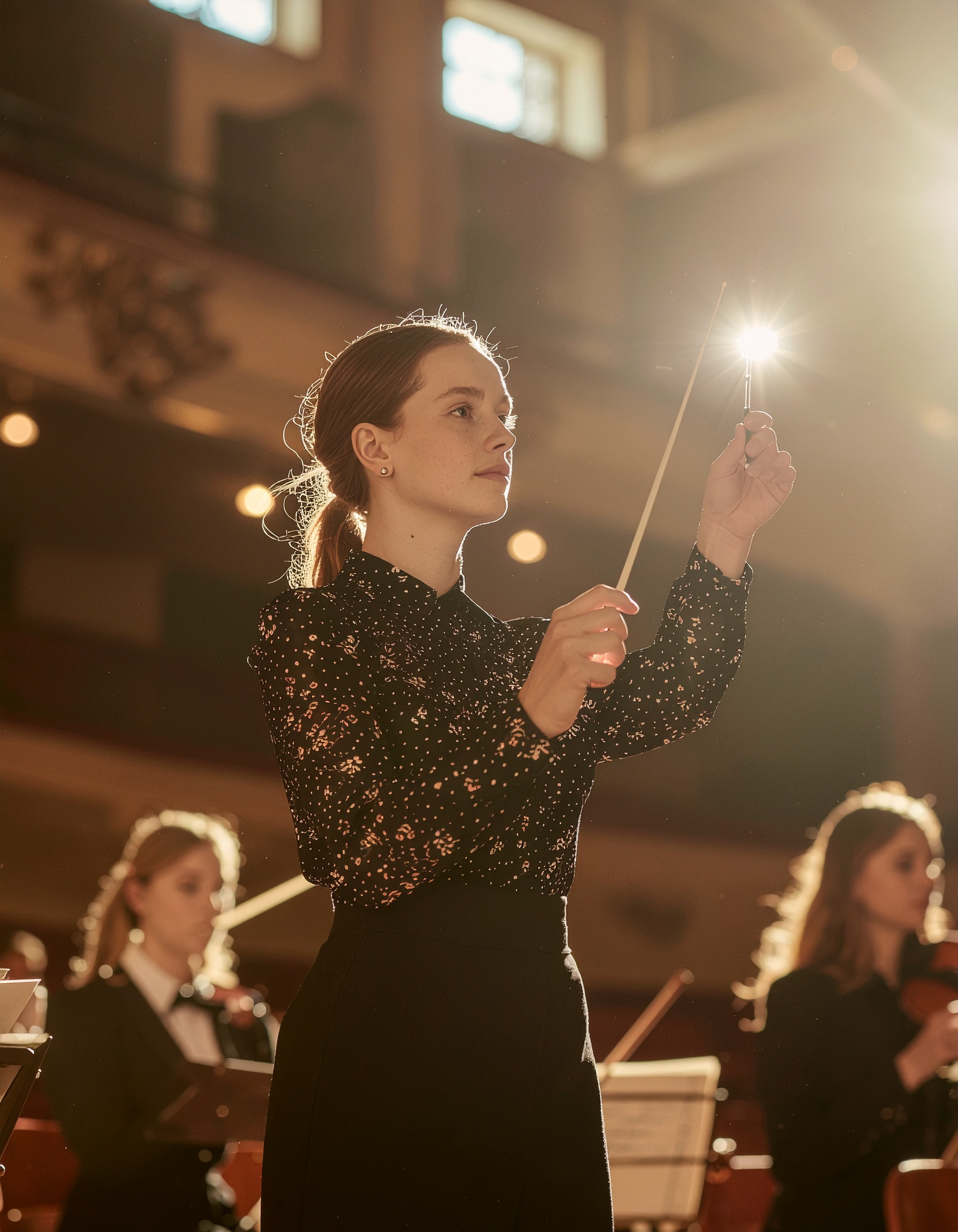 Uma maestrina jovem e confiante conduz uma orquestra em um palco iluminado por uma luz suave e quente. O foco da imagem está na expressão concentrada da maestrina e no brilho da batuta contra a iluminação teatral, criando uma atmosfera de dedicação e arte na música. A composição destaca também membros da orquestra ao fundo, misturando sombras e luzes para realçar a cena.