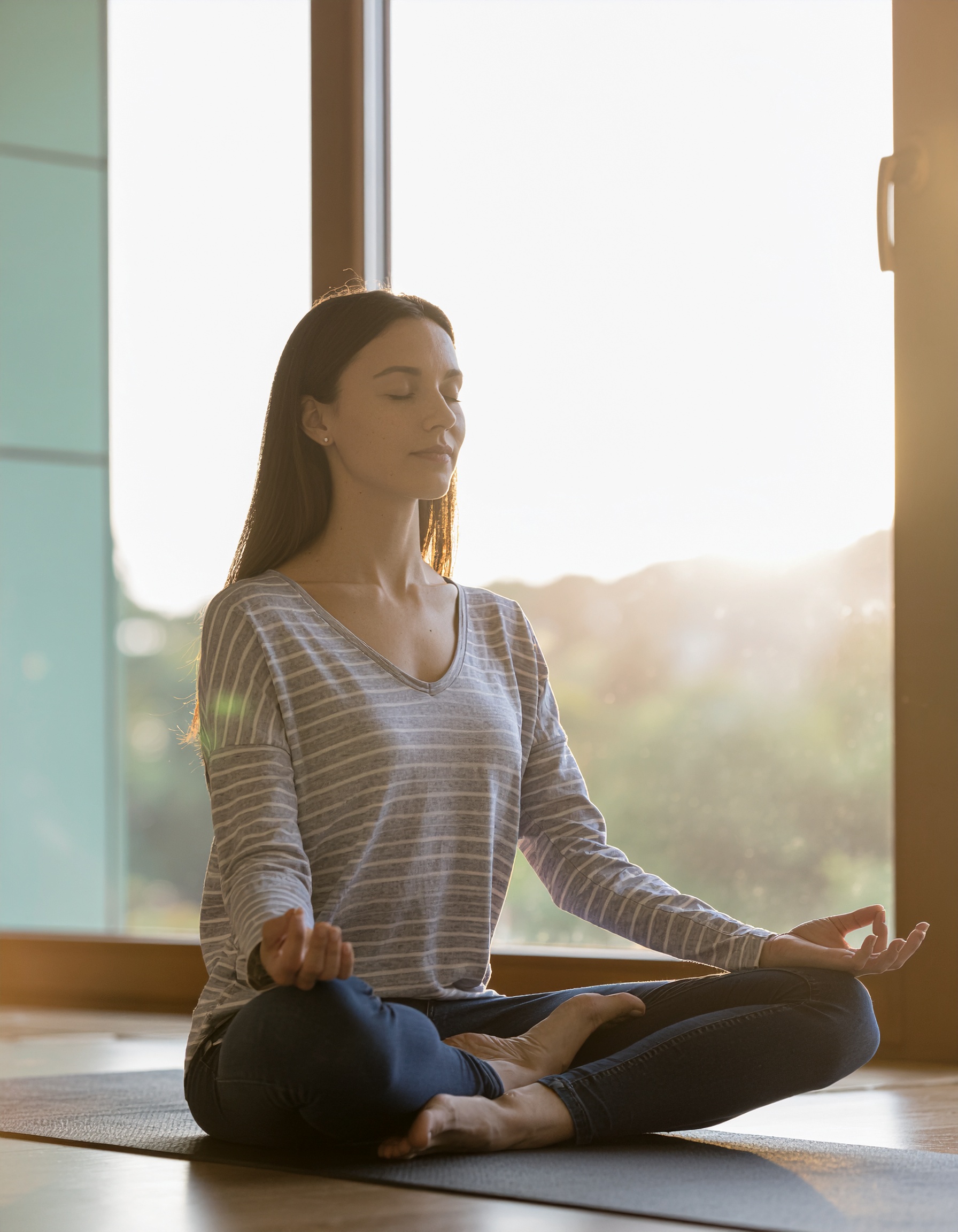Mulher sentada em posição de meditação em um ambiente interno iluminado pelo sol, usando camiseta listrada e calça jeans. A luz suave destaca a serenidade da cena, criando uma atmosfera de calma e introspecção. O foco está na postura relaxada e na expressão tranquila do rosto, com uma janela grande ao fundo permitindo a entrada de luz natural, acentuando a composição harmoniosa.
