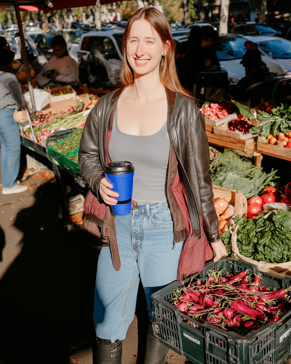 Mulher sorrindo segurando copo azul em feira ao ar livre, cercada por caixas de vegetais frescos sob luz solar intensa, atmosfera vibrante e acolhedora, foco nítido no rosto e produtos, composição equilibrada e cheia de cor.